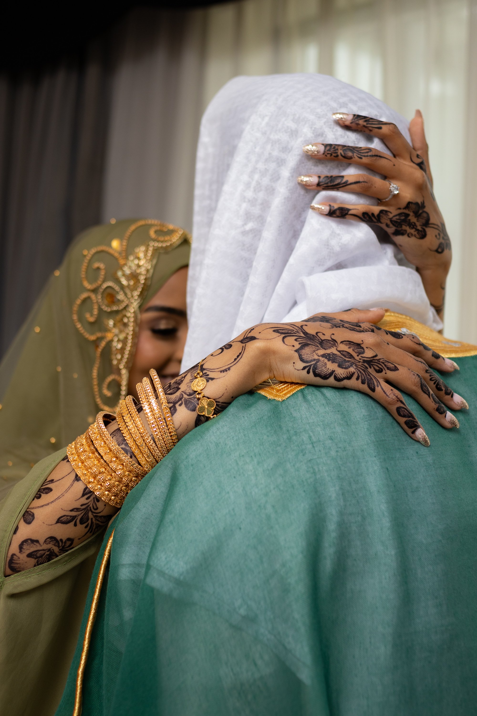 Close-up of two women hugging, with visible henna designs on their hands and arms. One woman is wearing a gold and green outfit with gold jewelry, and the other is dressed in white with decorative elements on their head covering. The background featu