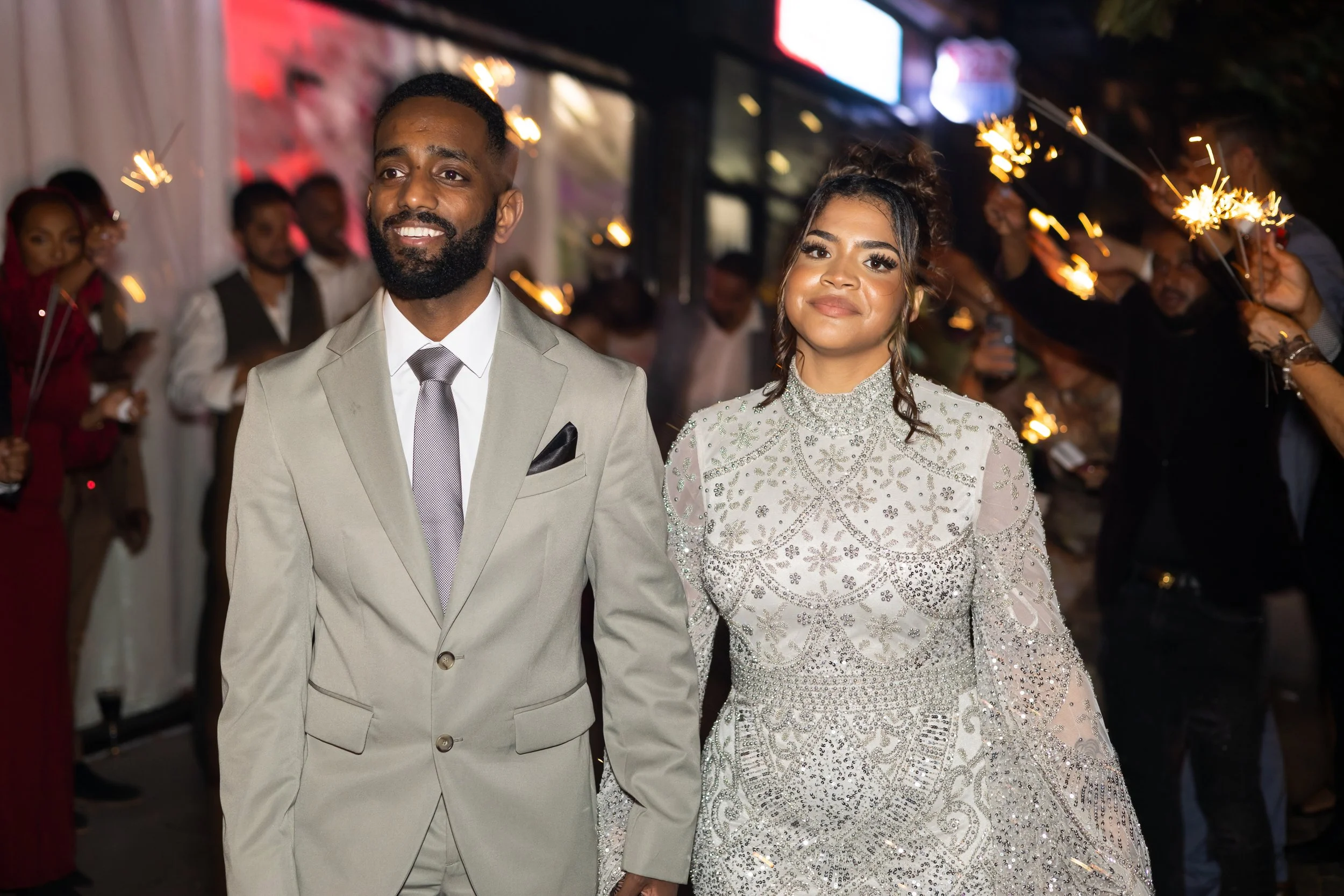 A man and woman dressed in formal attire celebrating at night, with sparklers and other party guests in the background.