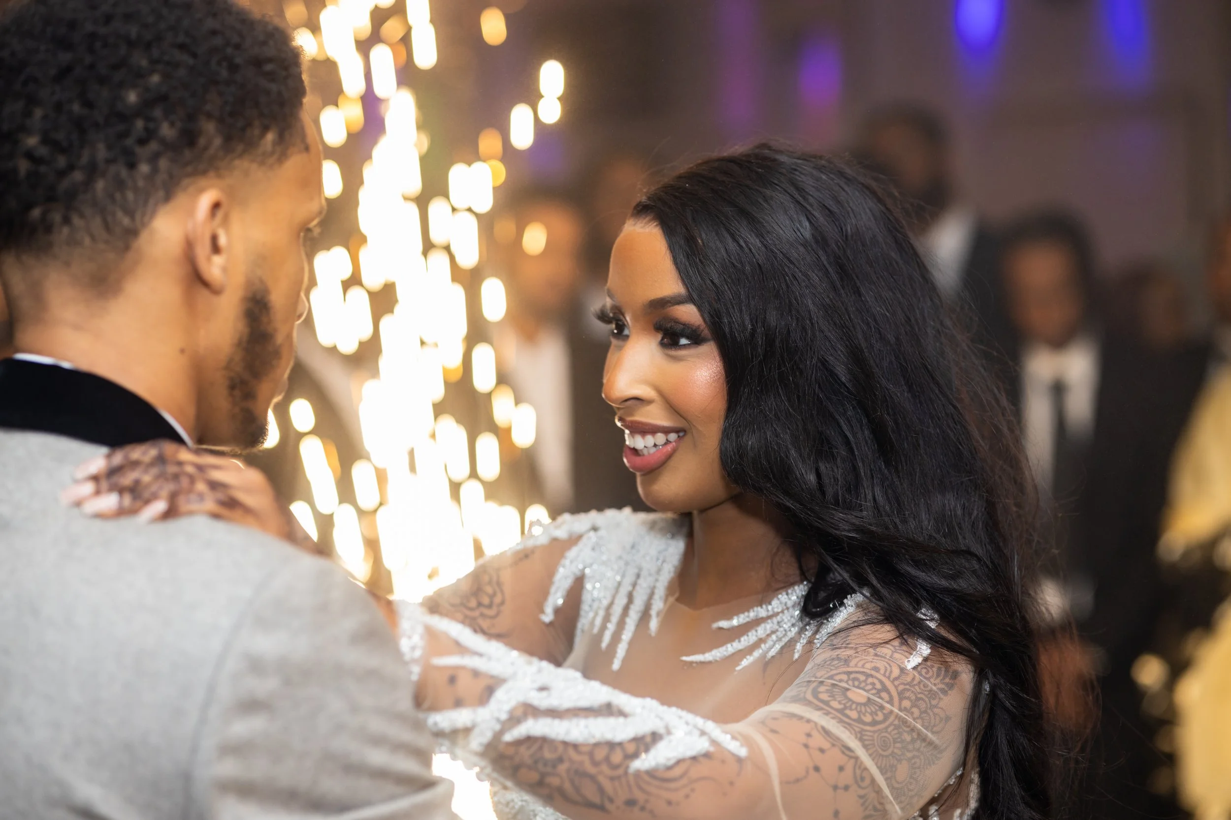 A couple dancing closely at a social event, with the woman smiling and showing tattoos on her arms, and the man facing her, with blurred background and warm lighting.