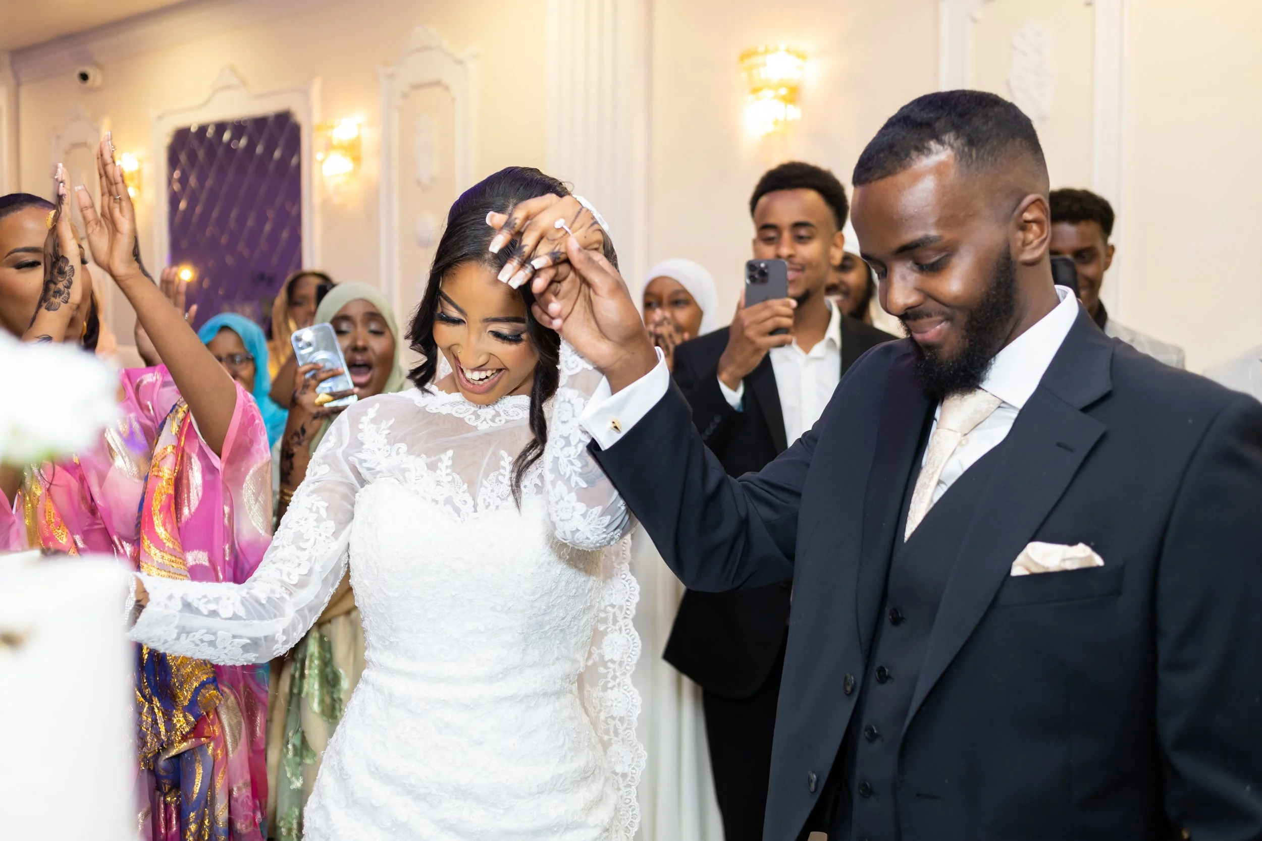 A bride and groom dancing at their wedding reception surrounded by friends and family, with some taking photos and videos.