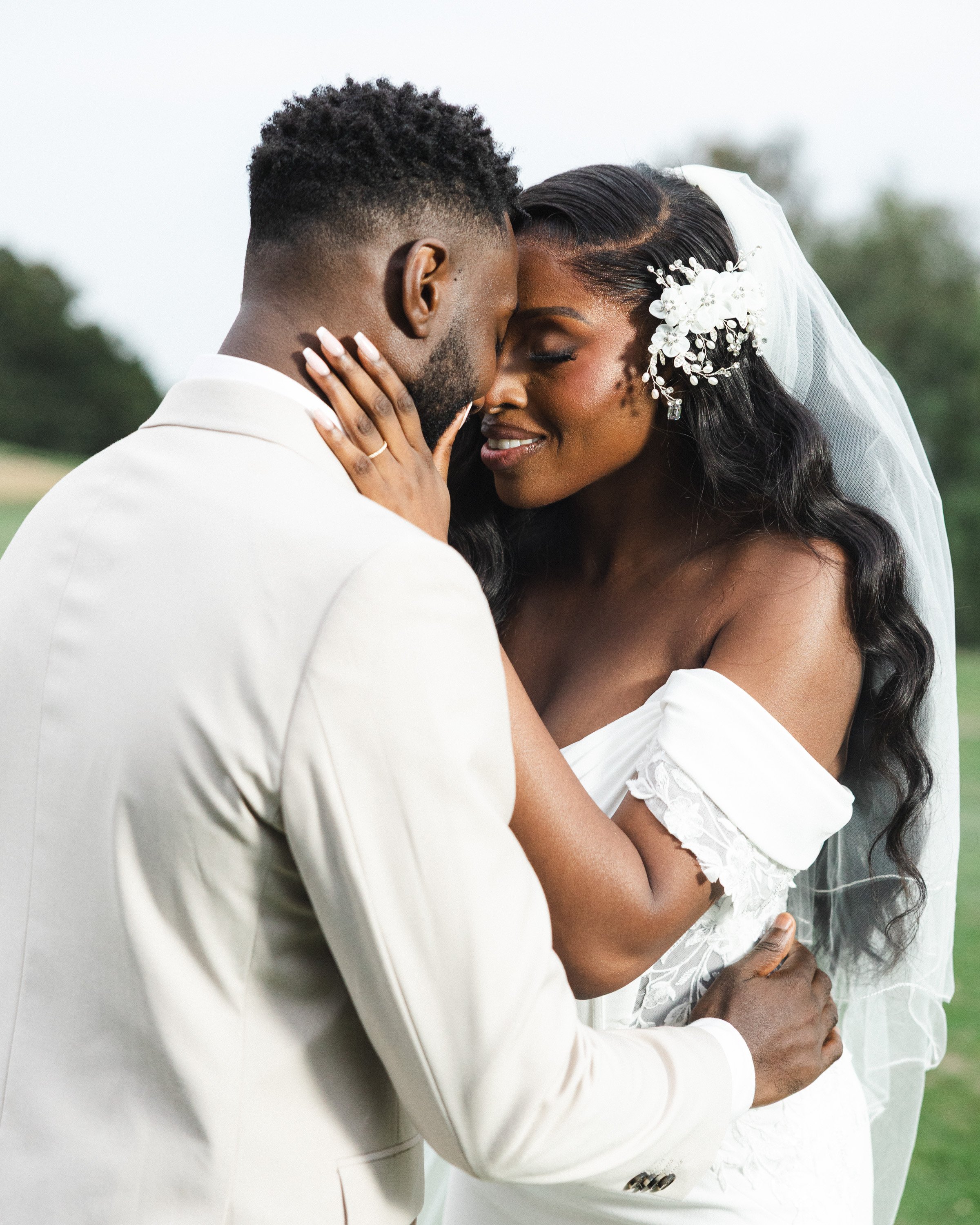 A groom and bride are holding each other and touching foreheads during their wedding ceremony outdoors.
