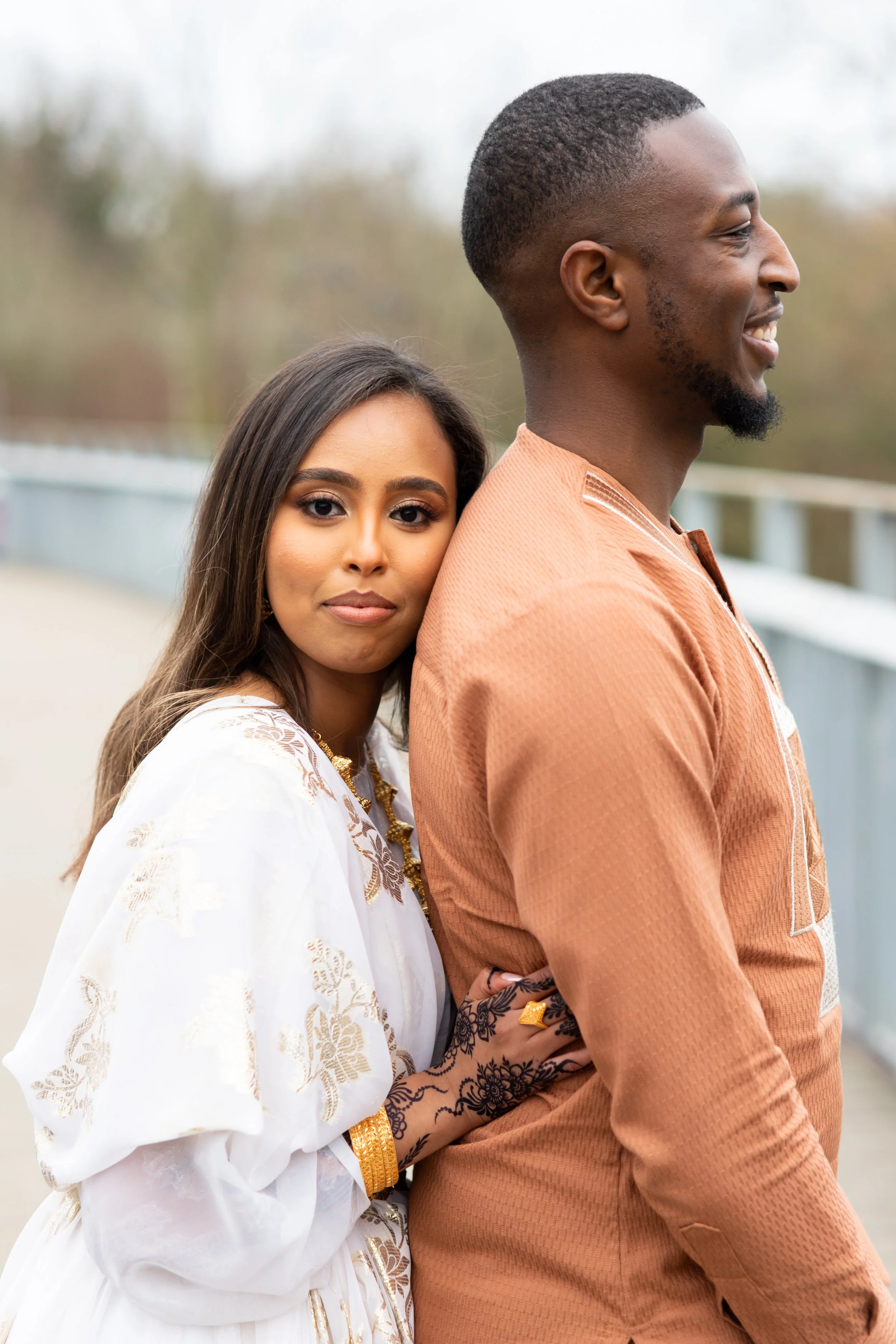 A woman with long brown hair and a man with a short haircut and a beard, standing outdoors. The woman is resting her head on the man's shoulder and holding his arm. The woman wears a white dress with gold embroidery, and the man wears an orange-brown