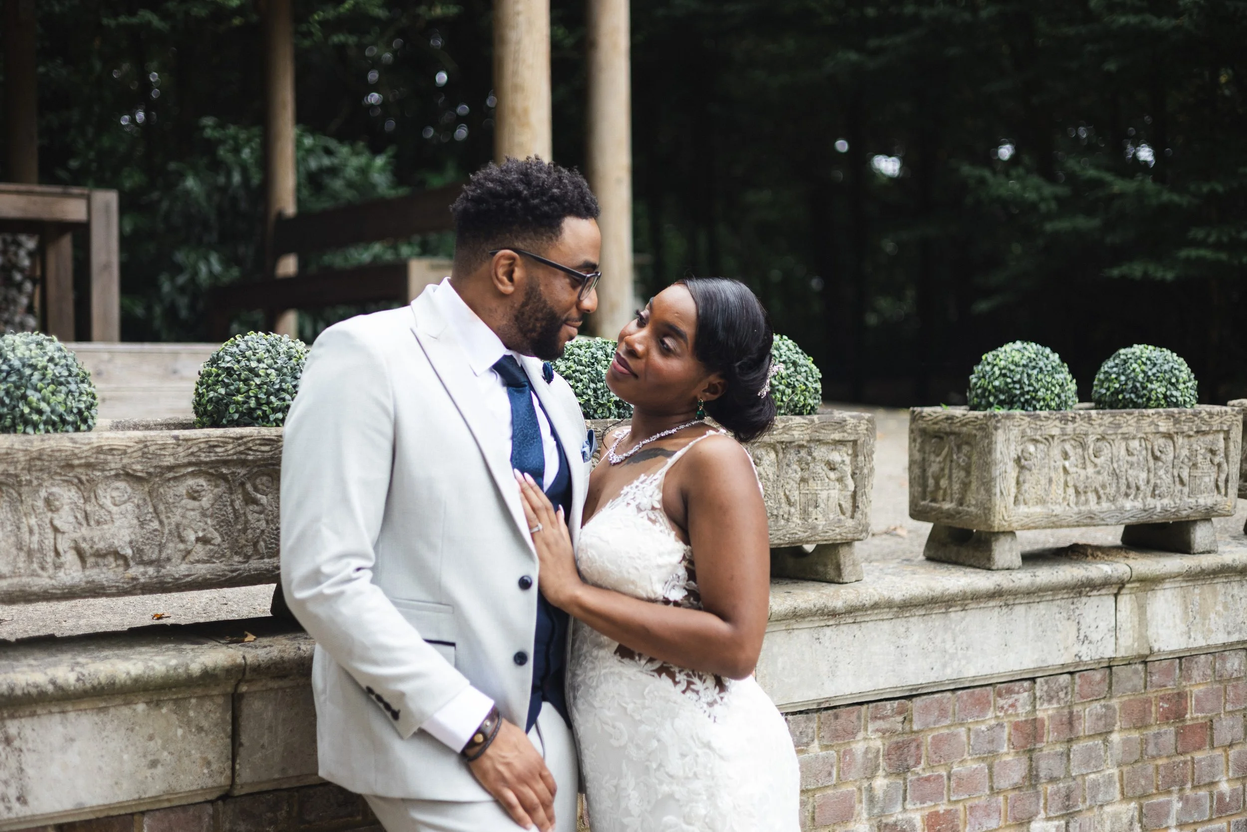 A bride and groom sharing an intimate moment outdoors, sitting on a stone bench with decorative stone planters behind them, surrounded by trees.