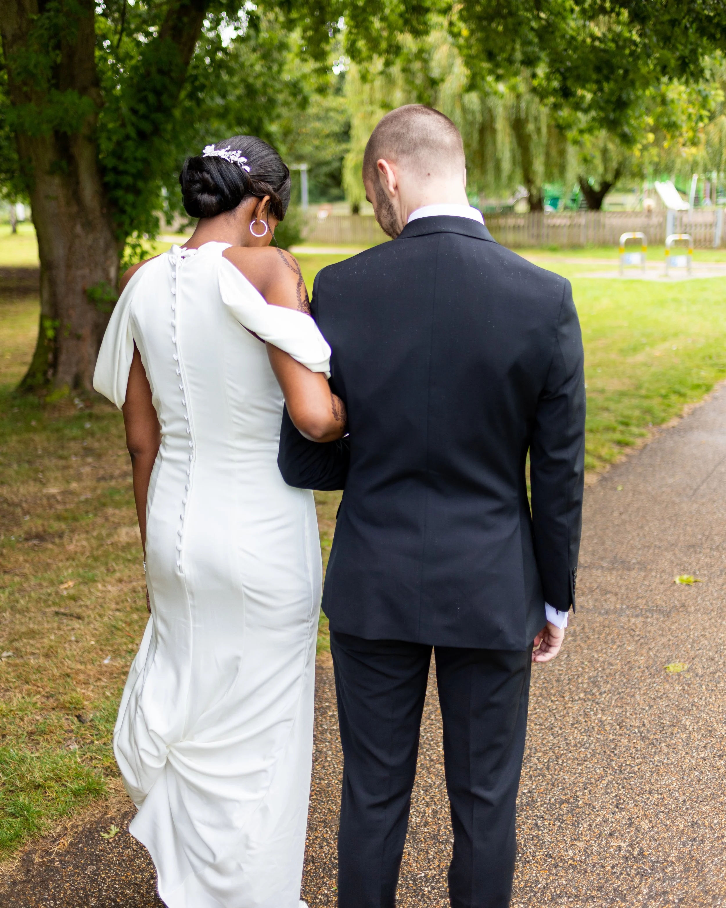 A bride and groom are walking together outdoors on a gravel path surrounded by green trees, seen from behind.