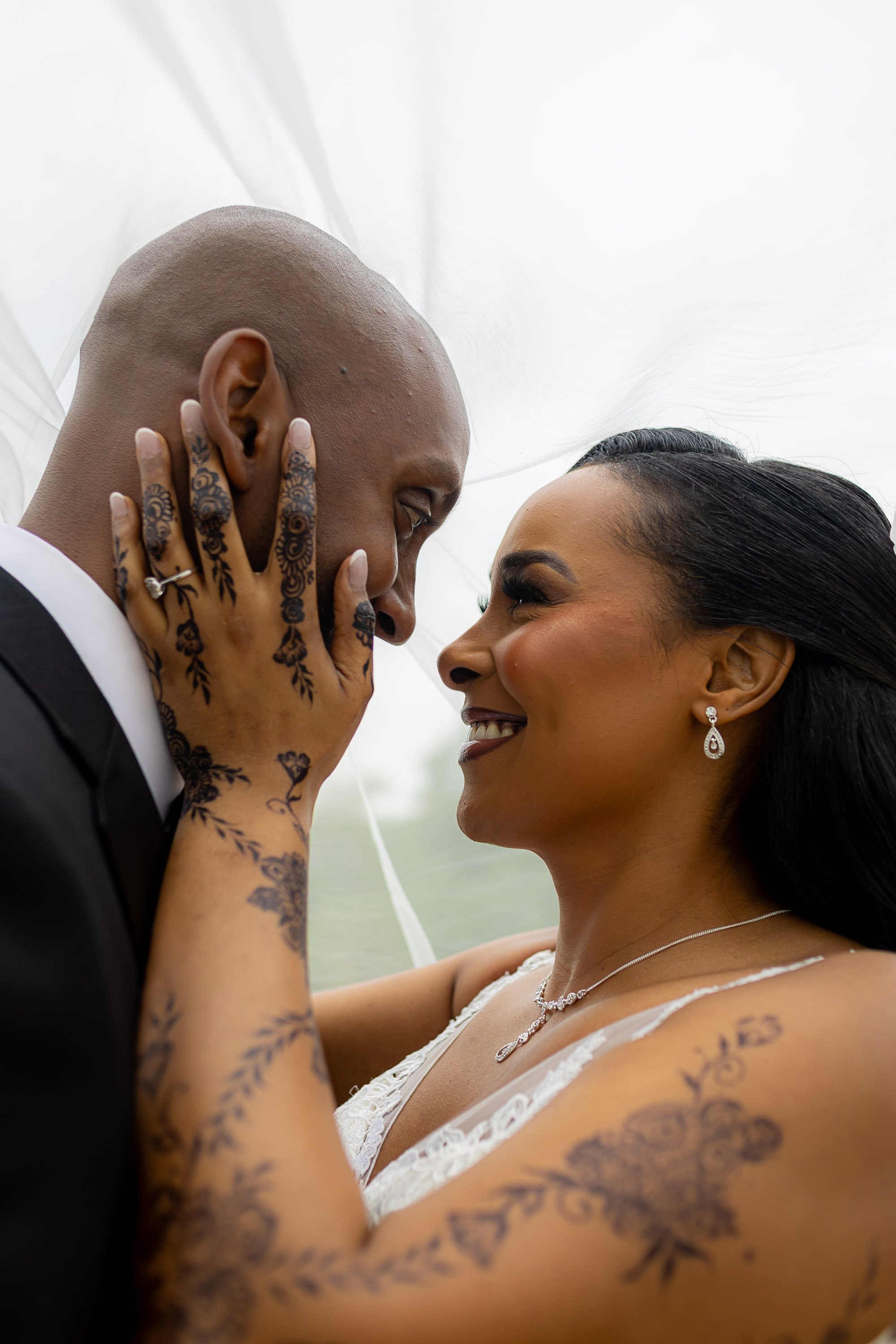 A wedding couple is close together with their foreheads touching, smiling warmly at each other. The bride is touching the groom's face and hand, revealing tattoos on her hand and arm.