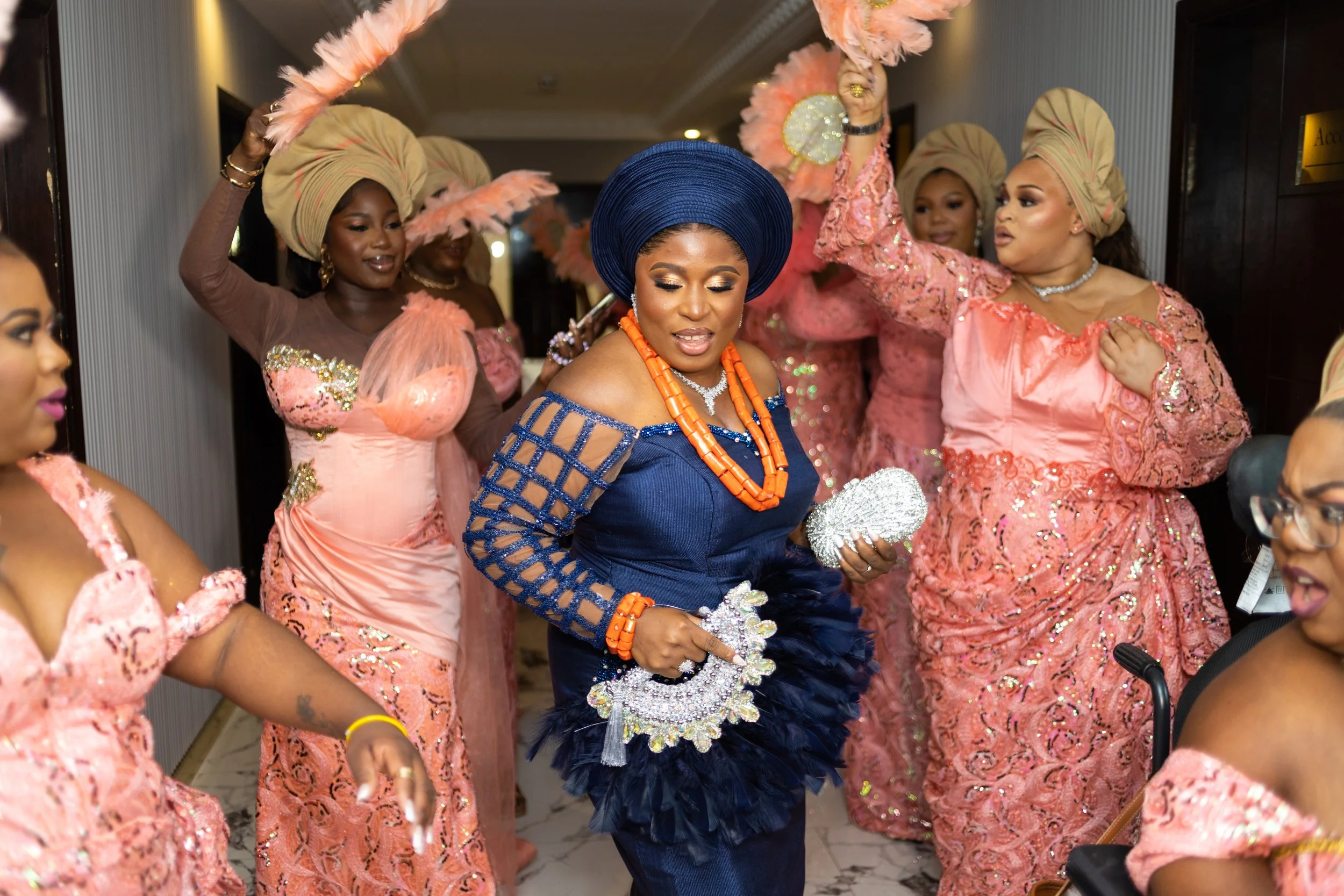 Women dressed in traditional and elegant clothing, including gele head wraps, dancing and celebrating at a cultural event or ceremony.