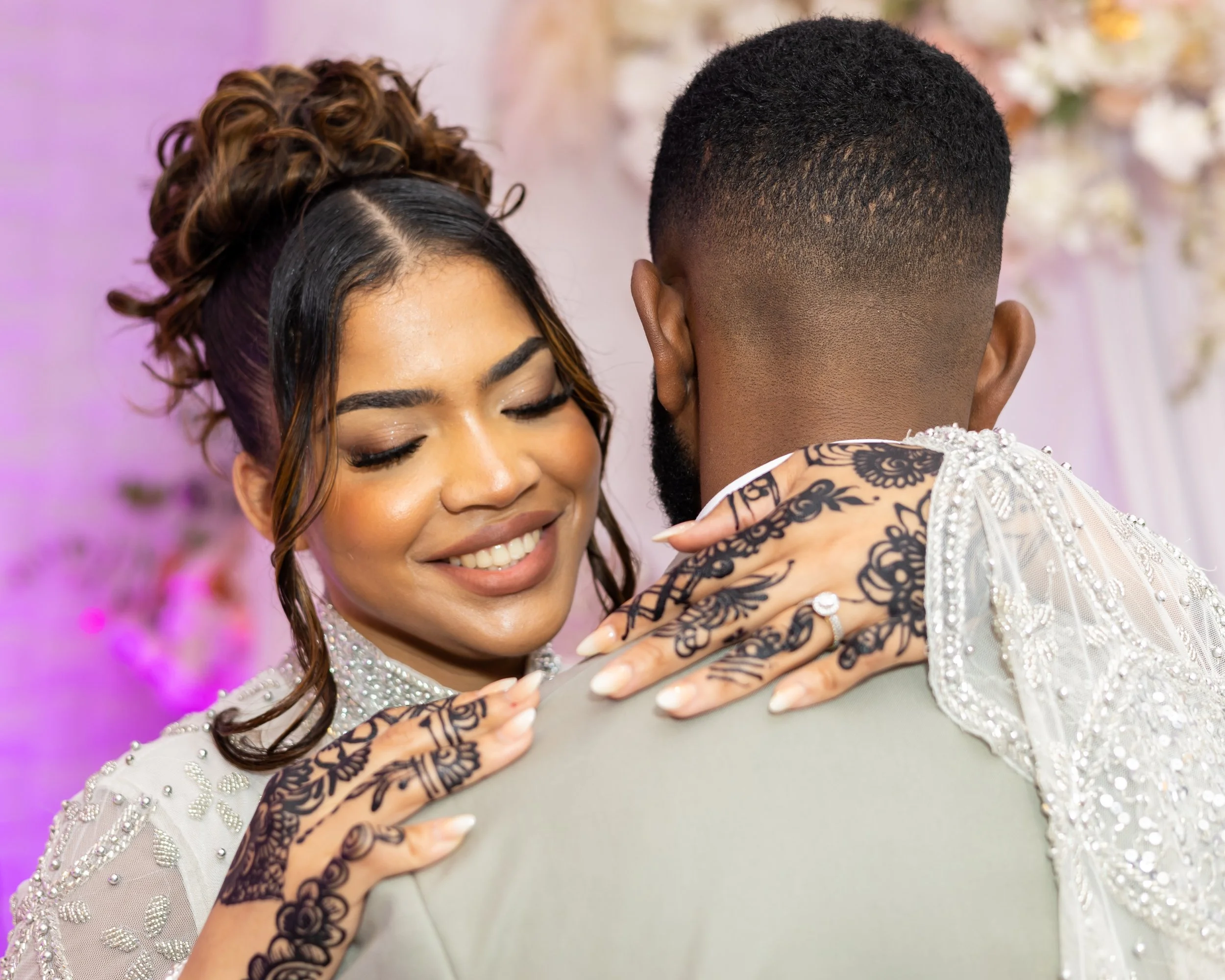 Close-up of a couple dancing, with the woman smiling and the man facing away, revealing a tattoo on his arm, both wearing wedding attire.
