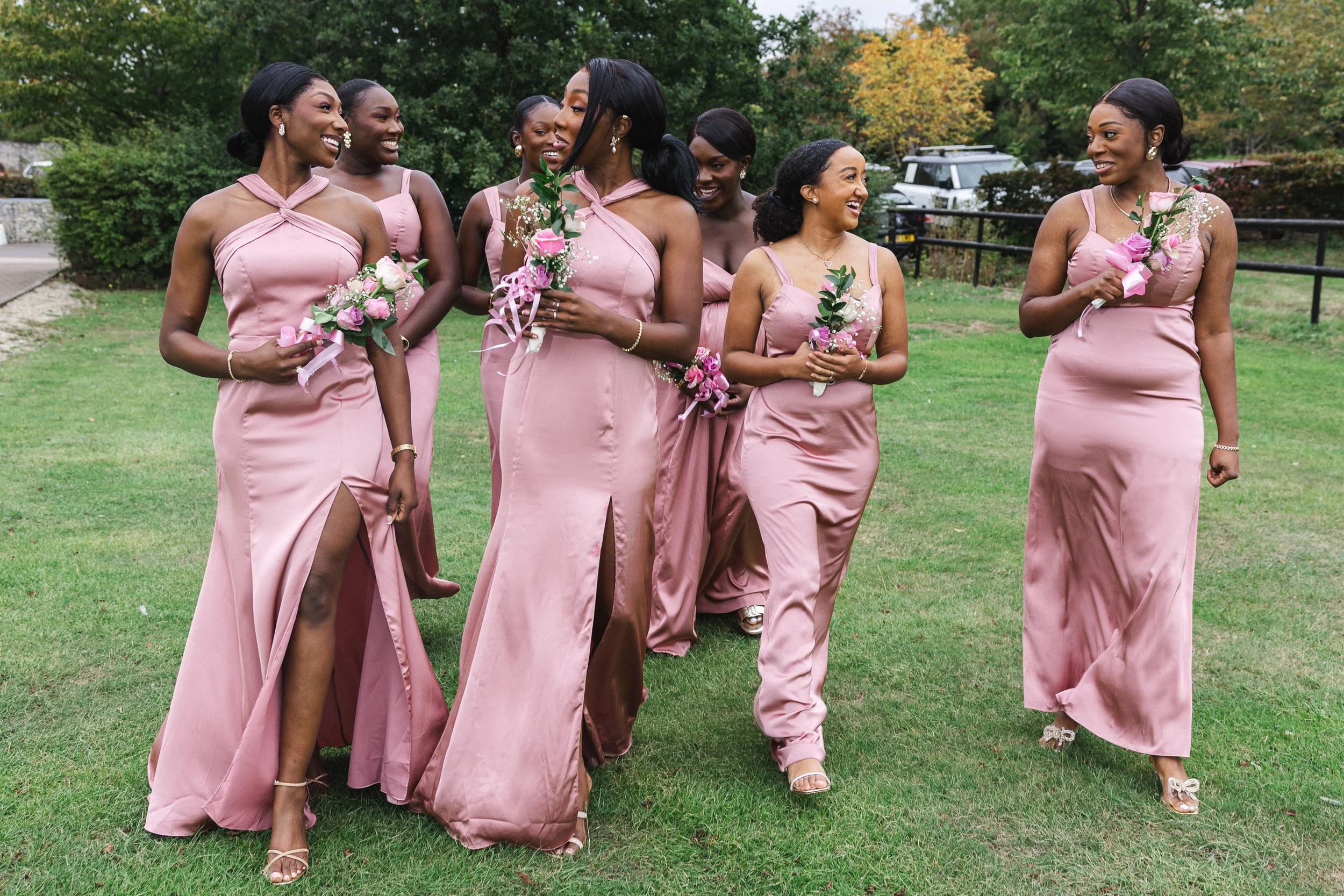 Group of women in matching pink dresses holding bouquets, walking on a grassy outdoor area with trees in the background.