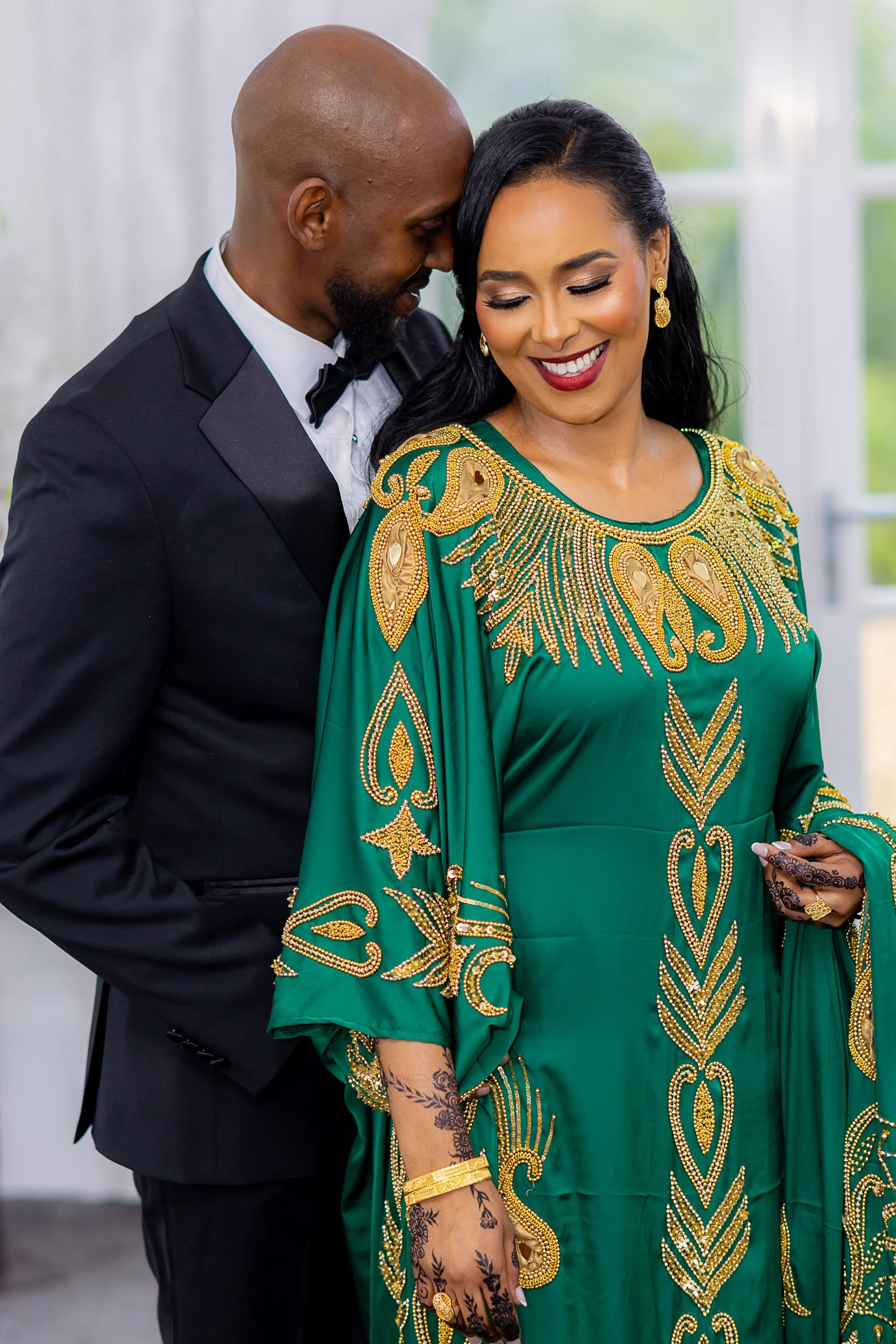 A man in a black tuxedo with a bow tie and a woman in a green gown with gold embroidery, smiling and touching foreheads in an intimate moment indoors with a blurred window in the background.