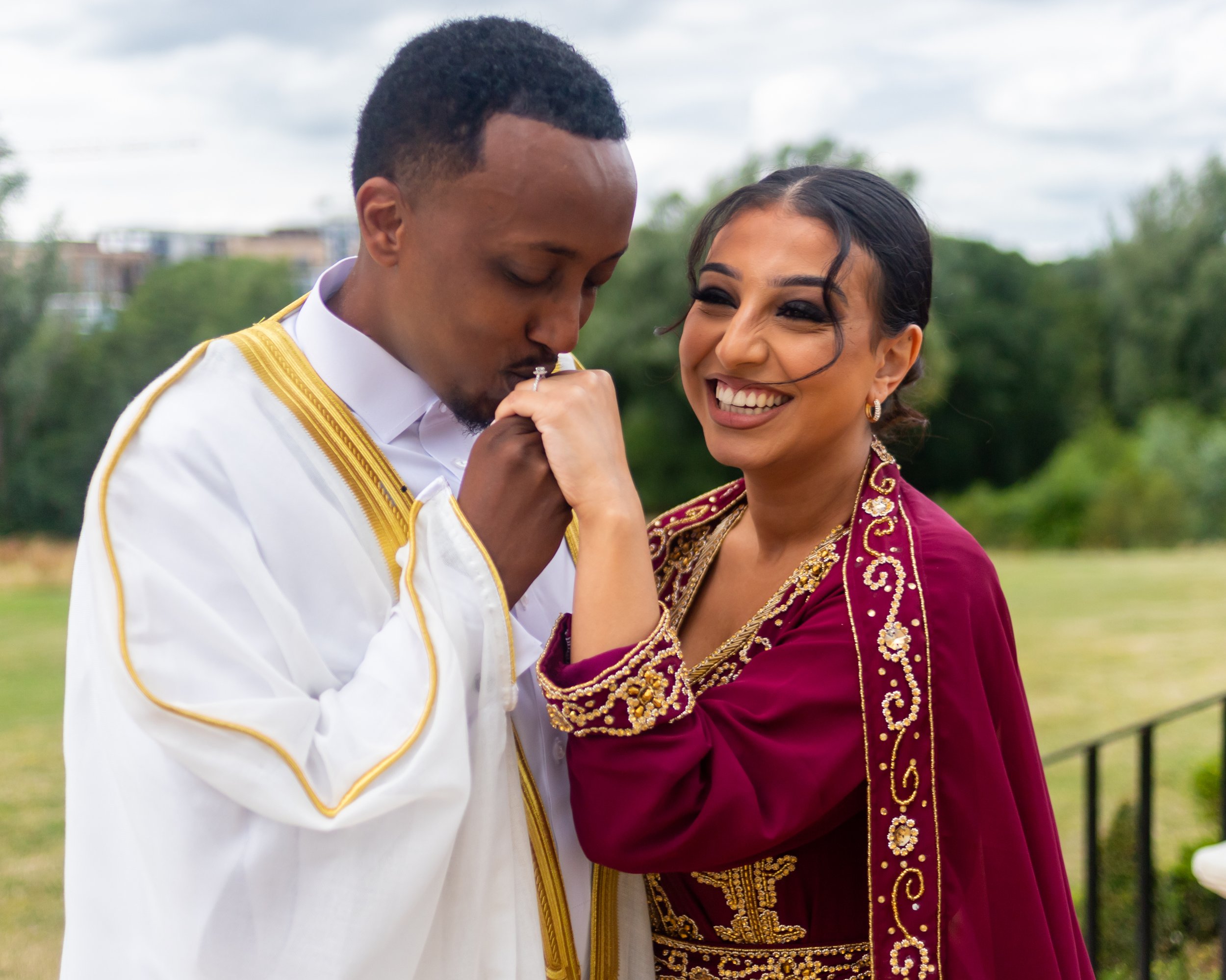 A couple dressed in formal attire, the man in a white suit with gold trim and the woman in a burgundy and gold embroidered dress, sharing an intimate moment outdoors with a cloudy sky and greenery in the background.