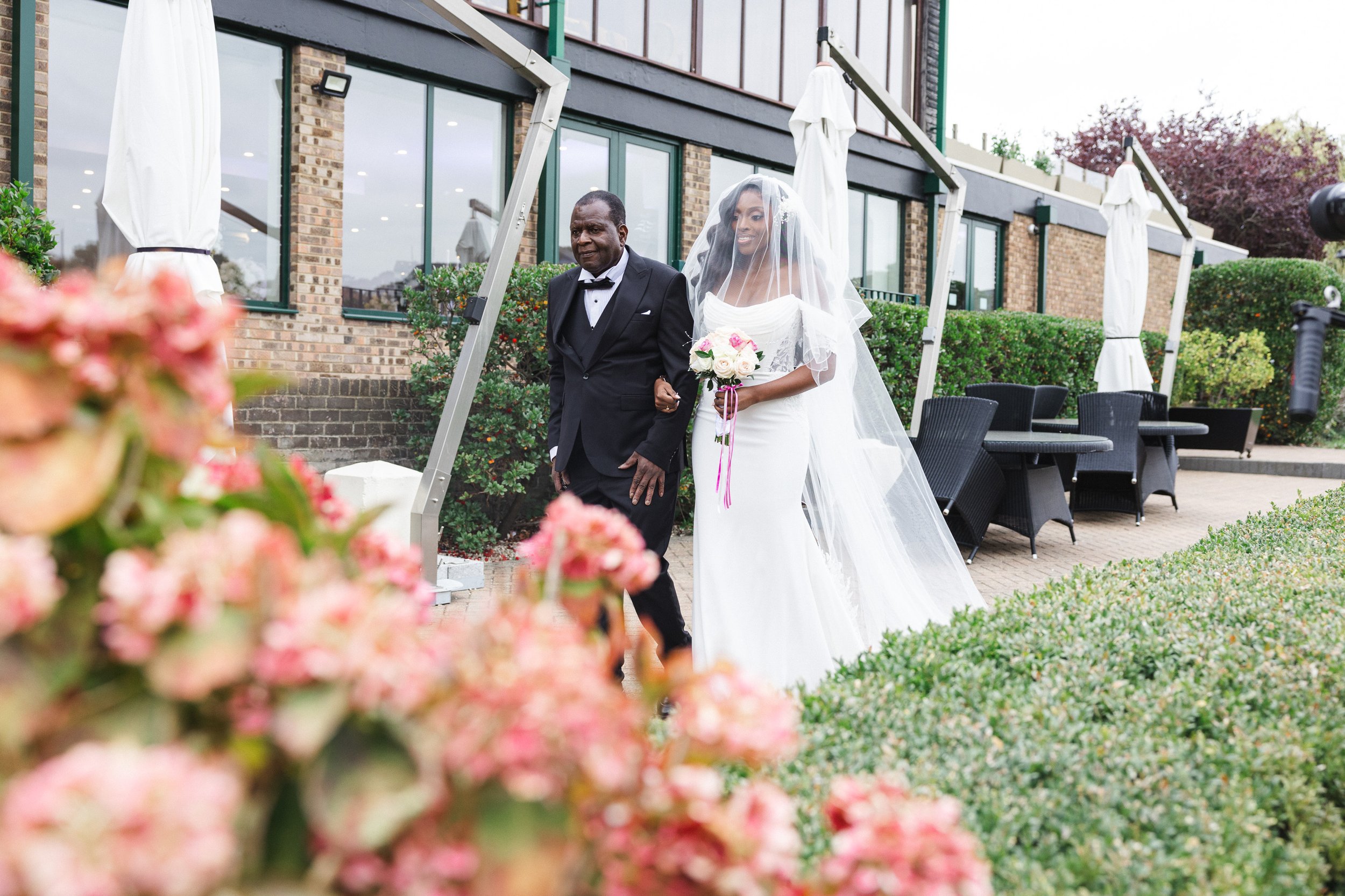 A bride and an older man walking outdoors near a brick building with large windows, surrounded by floral bushes and outdoor furniture, during a wedding or special event.