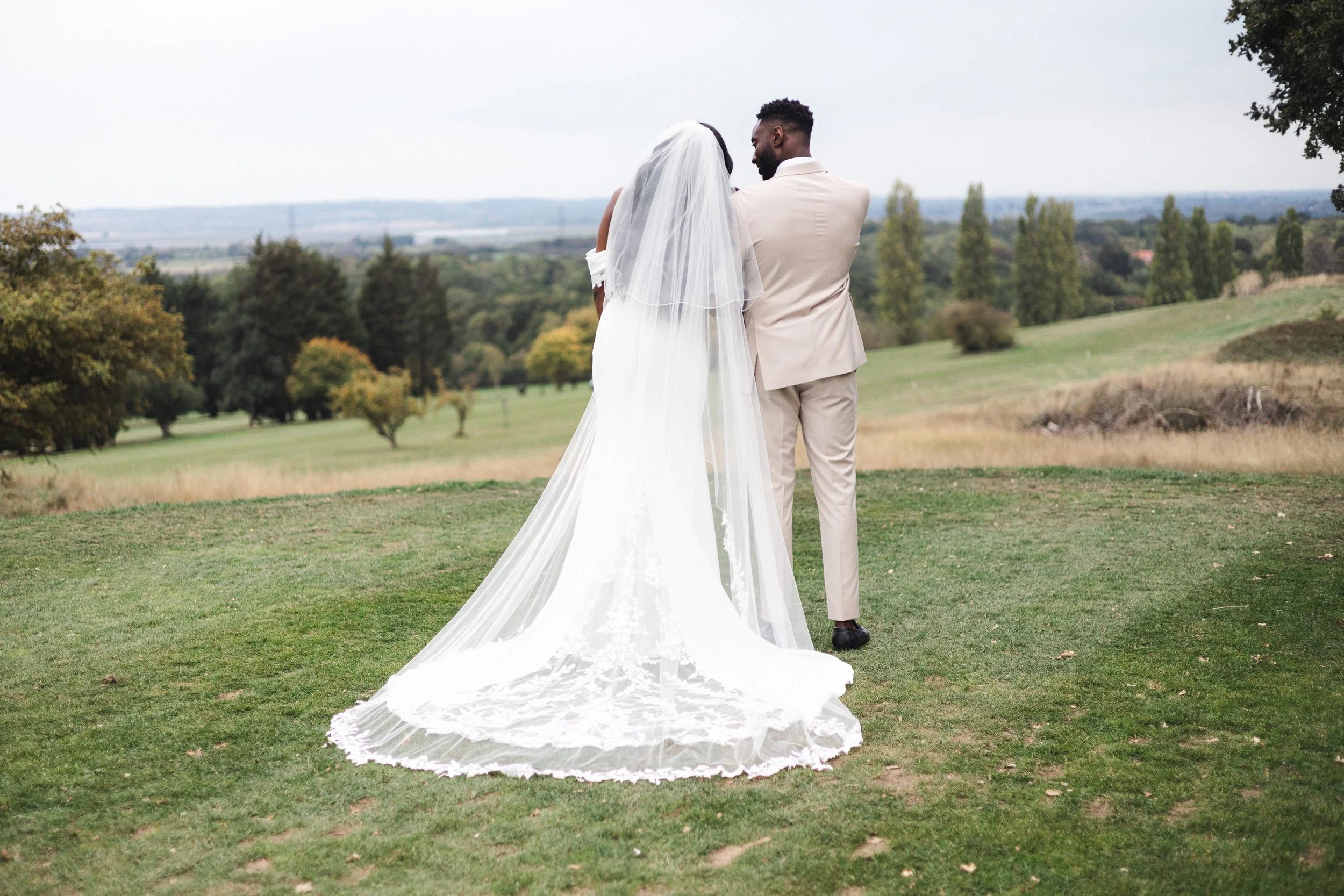 A bride and groom stand together on a grassy field, with the bride wearing a long white wedding dress and veil, and the groom in a light-colored suit. They are facing away from the camera, looking at the landscape of trees and rolling hills in the di