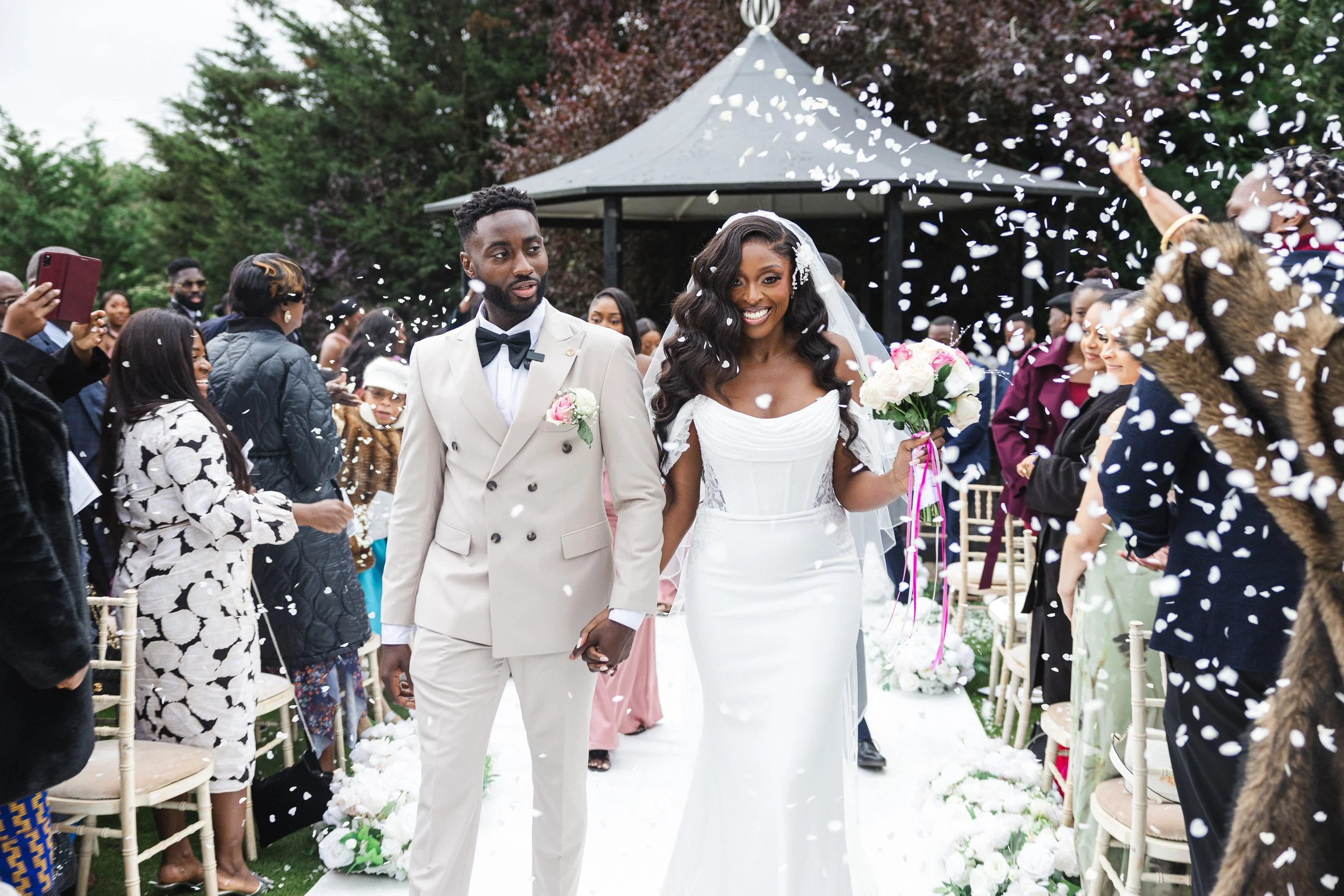 A bride and groom are walking hand in hand down an outdoor wedding aisle, surrounded by guests throwing flower petals, under a canopy, with trees in the background.