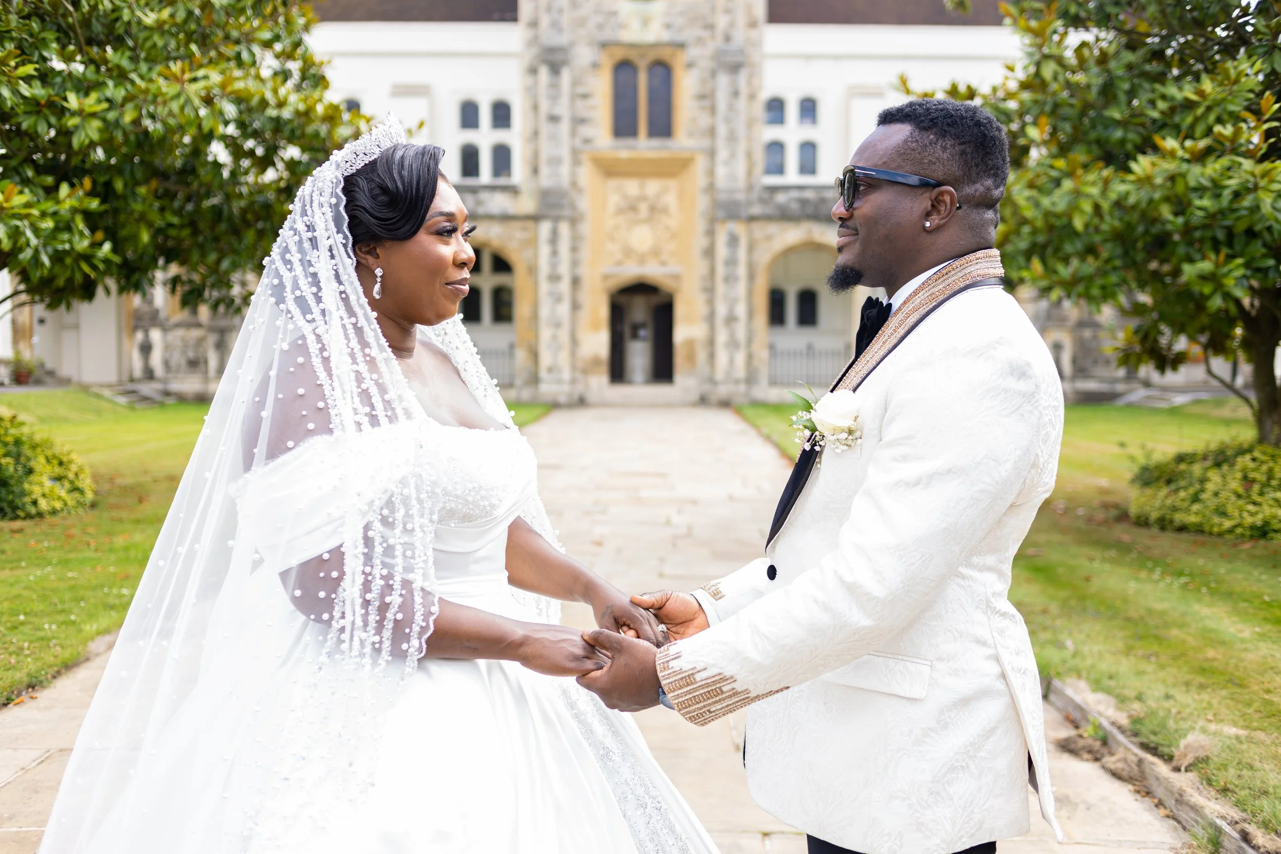 A bride and groom holding hands during their wedding ceremony outdoors, standing in front of a historic building with greenery around.