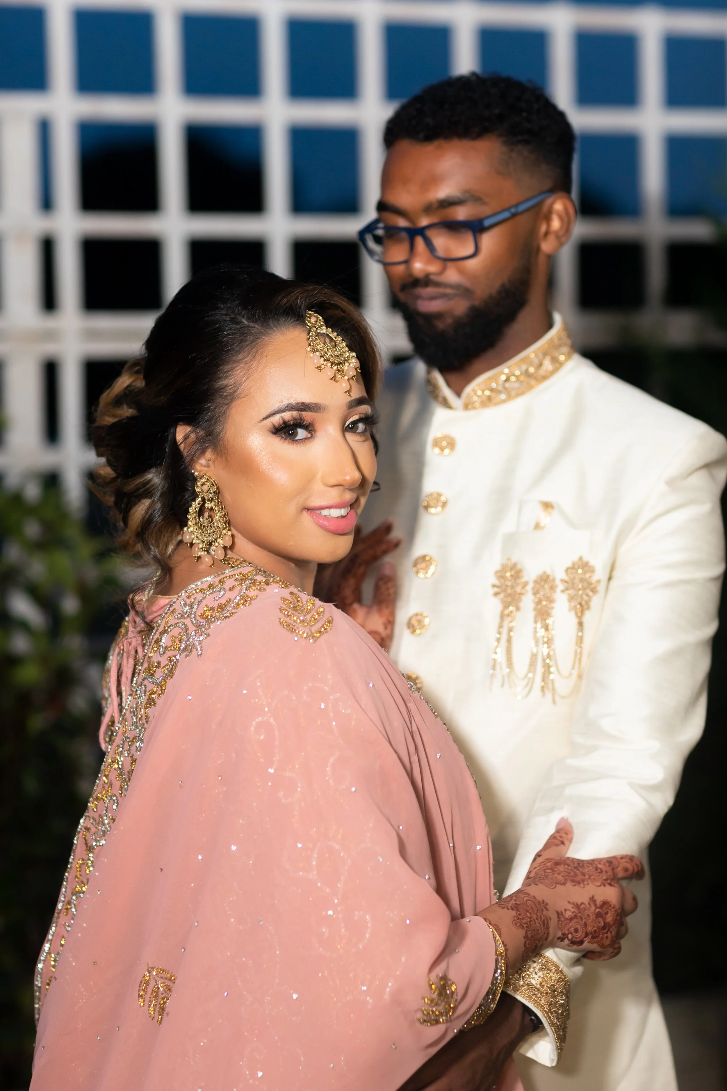 A Indian bride and groom dressed in traditional attire, with the bride wearing a pink and gold embroidered saree and the groom in a white sherwani with gold accents, stand close together.