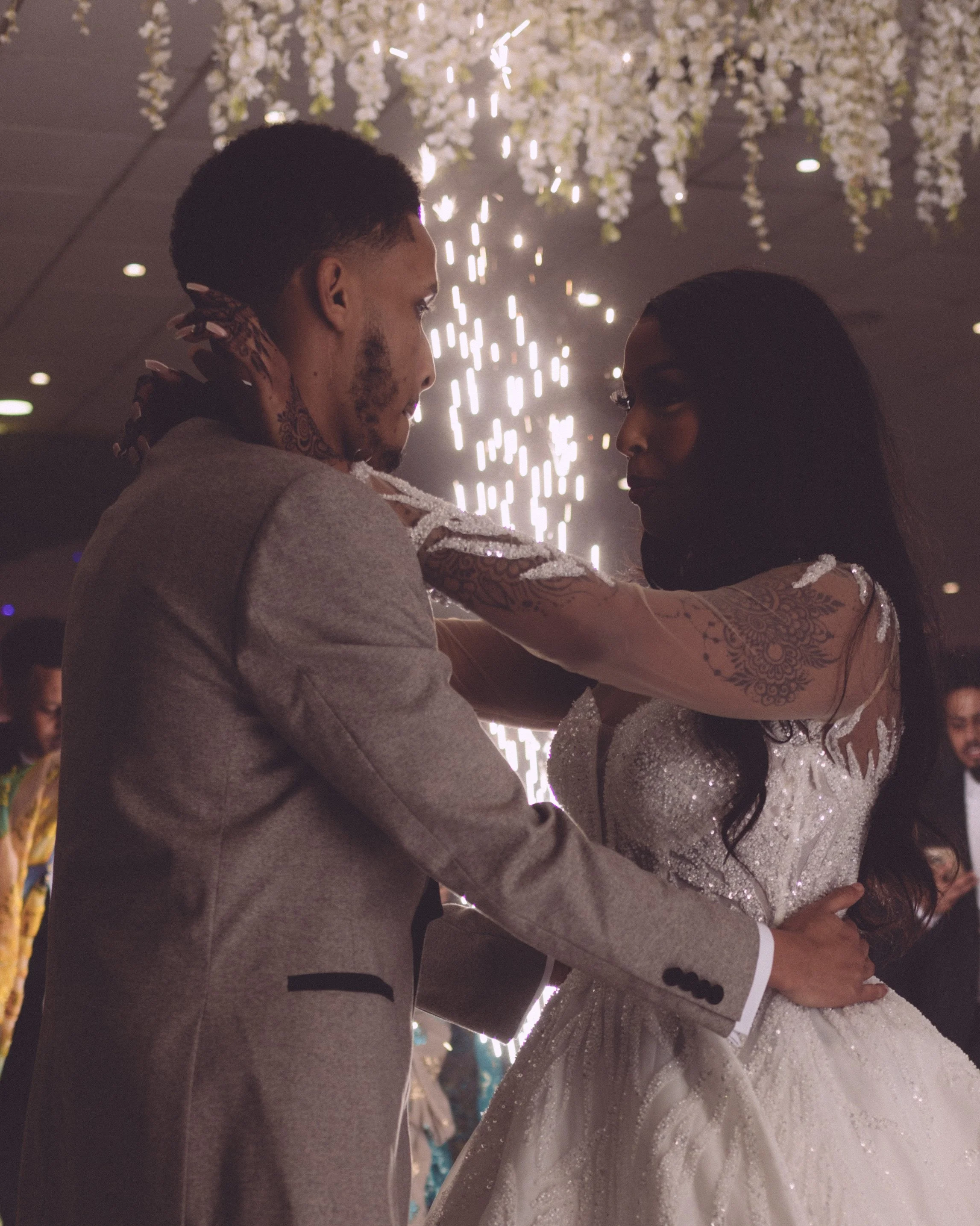 A couple is dancing closely at their wedding reception, with the groom in a grey suit and the bride in a white, sequined wedding gown. They are holding each other and looking into each other's eyes under a ceiling decorated with hanging white flowers