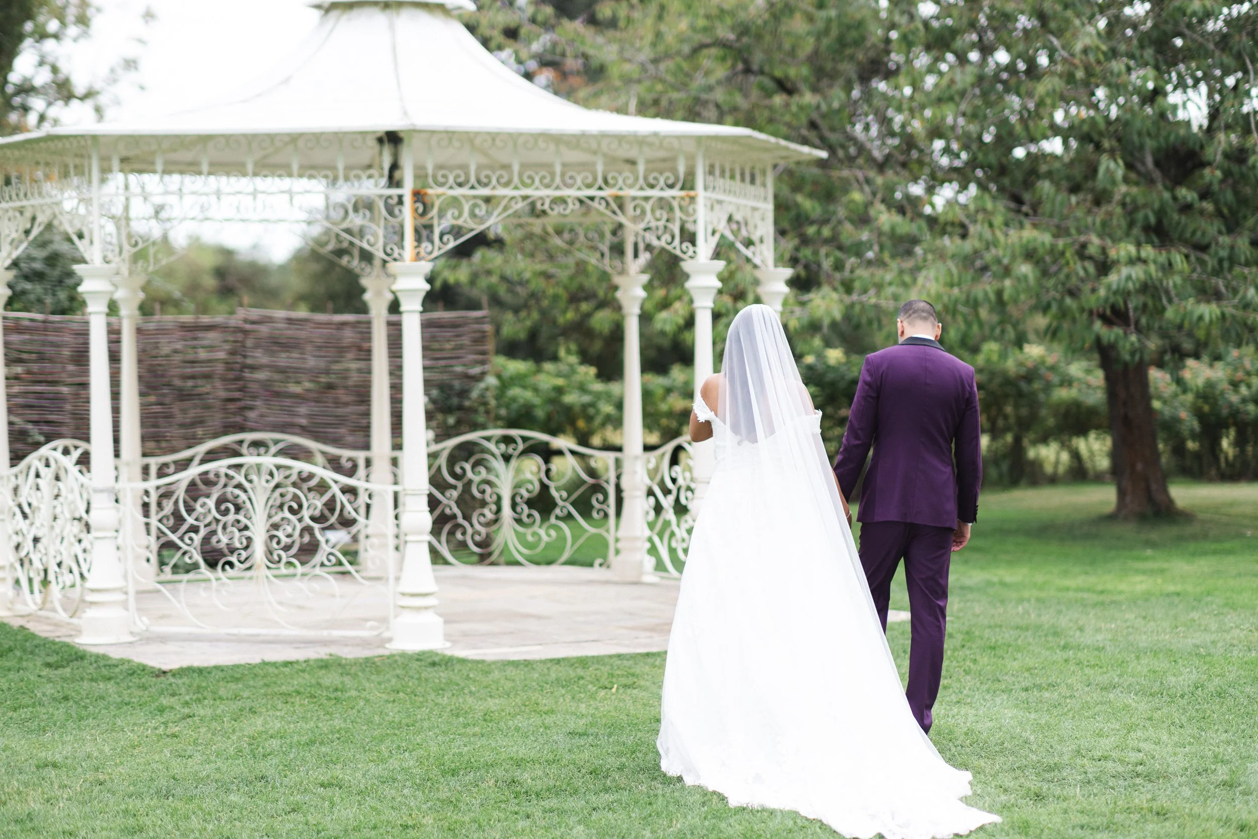 A bride and groom walking away from a white ornate gazebo in a garden, with trees and grass surrounding them during outdoor wedding.