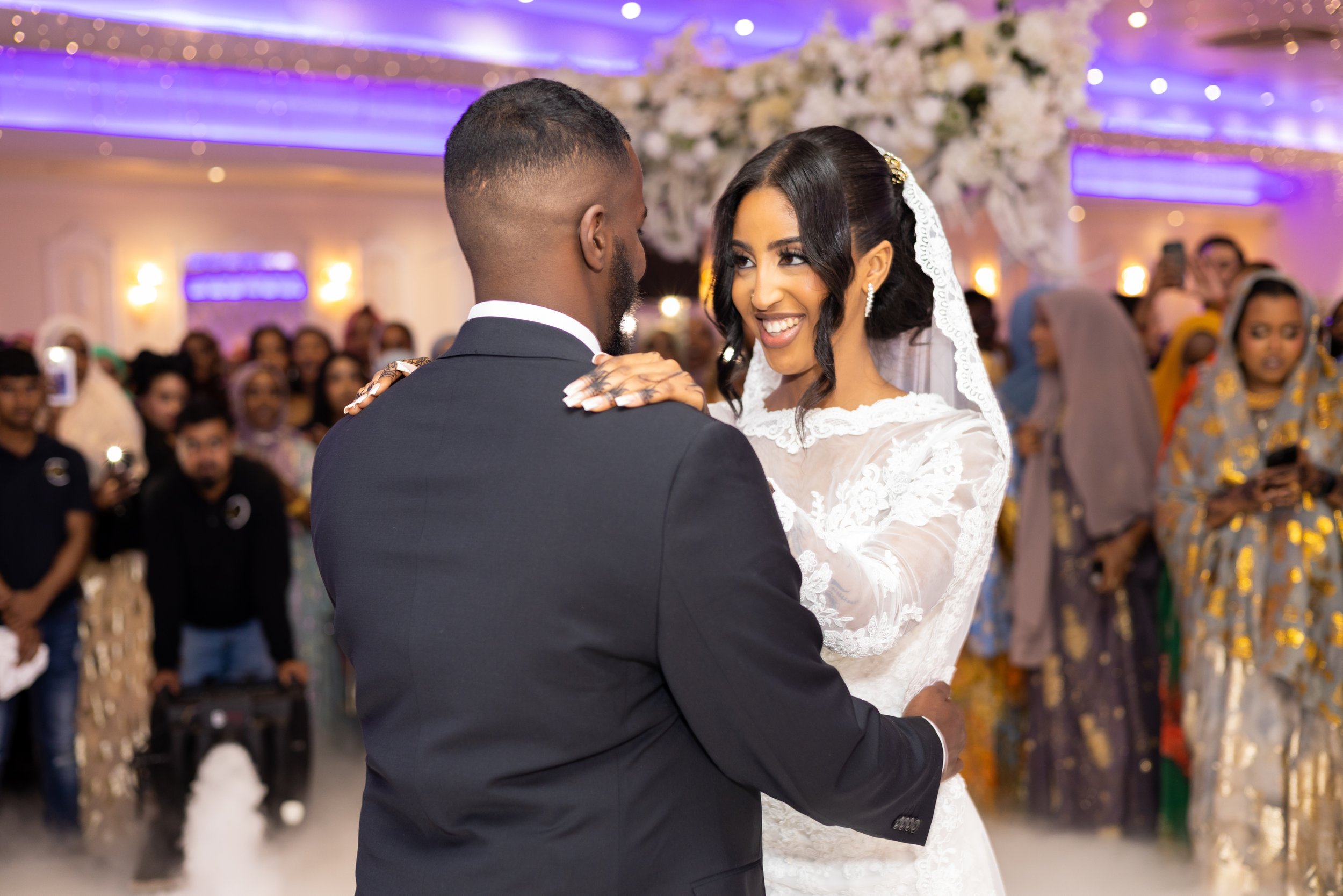 A bride and groom sharing a dance at their wedding reception, surrounded by guests in a decorated hall with floral arrangements and colorful lighting.