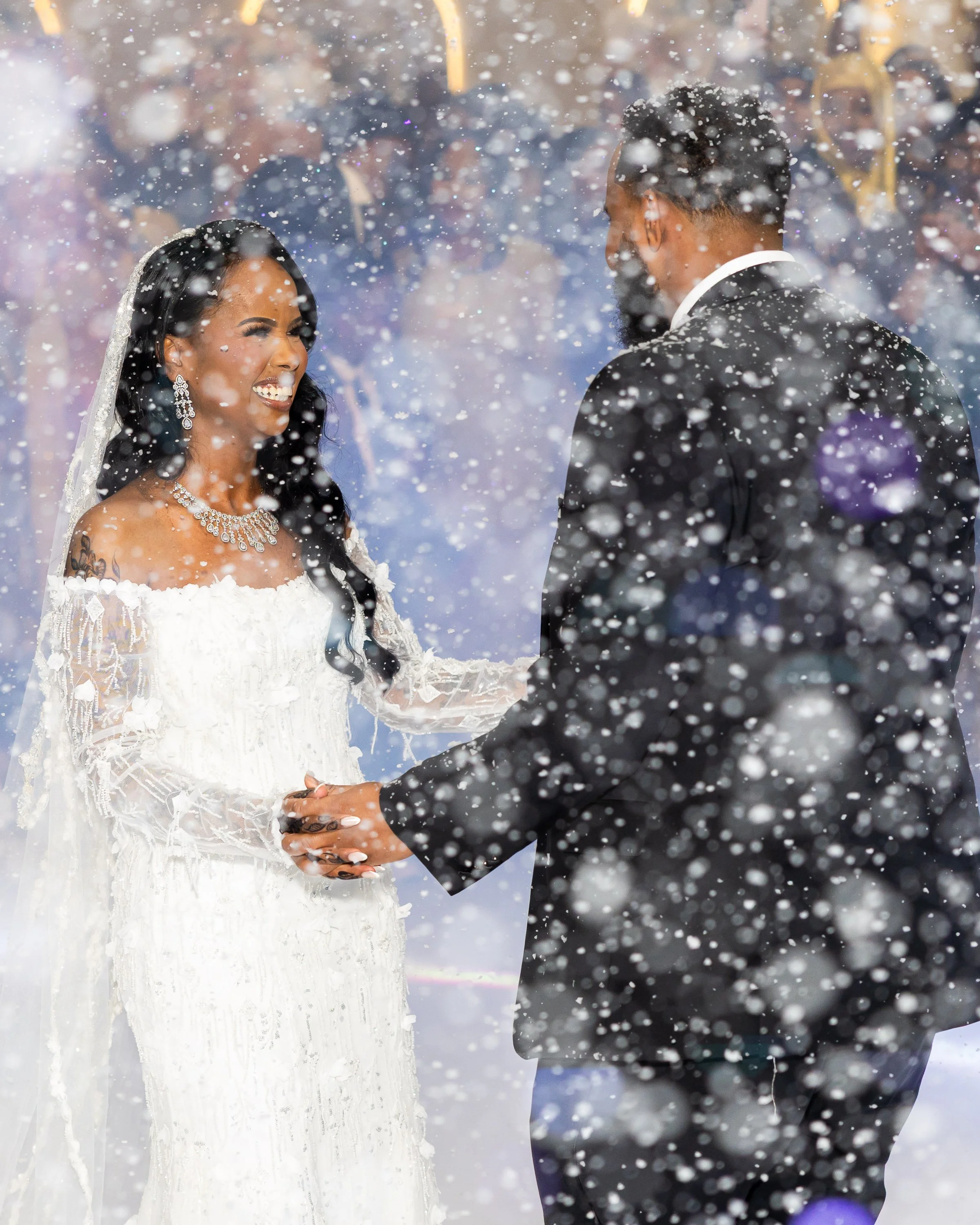 A bride and groom hold hands during their wedding ceremony in a snowy setting, with snow falling and guests in the background.