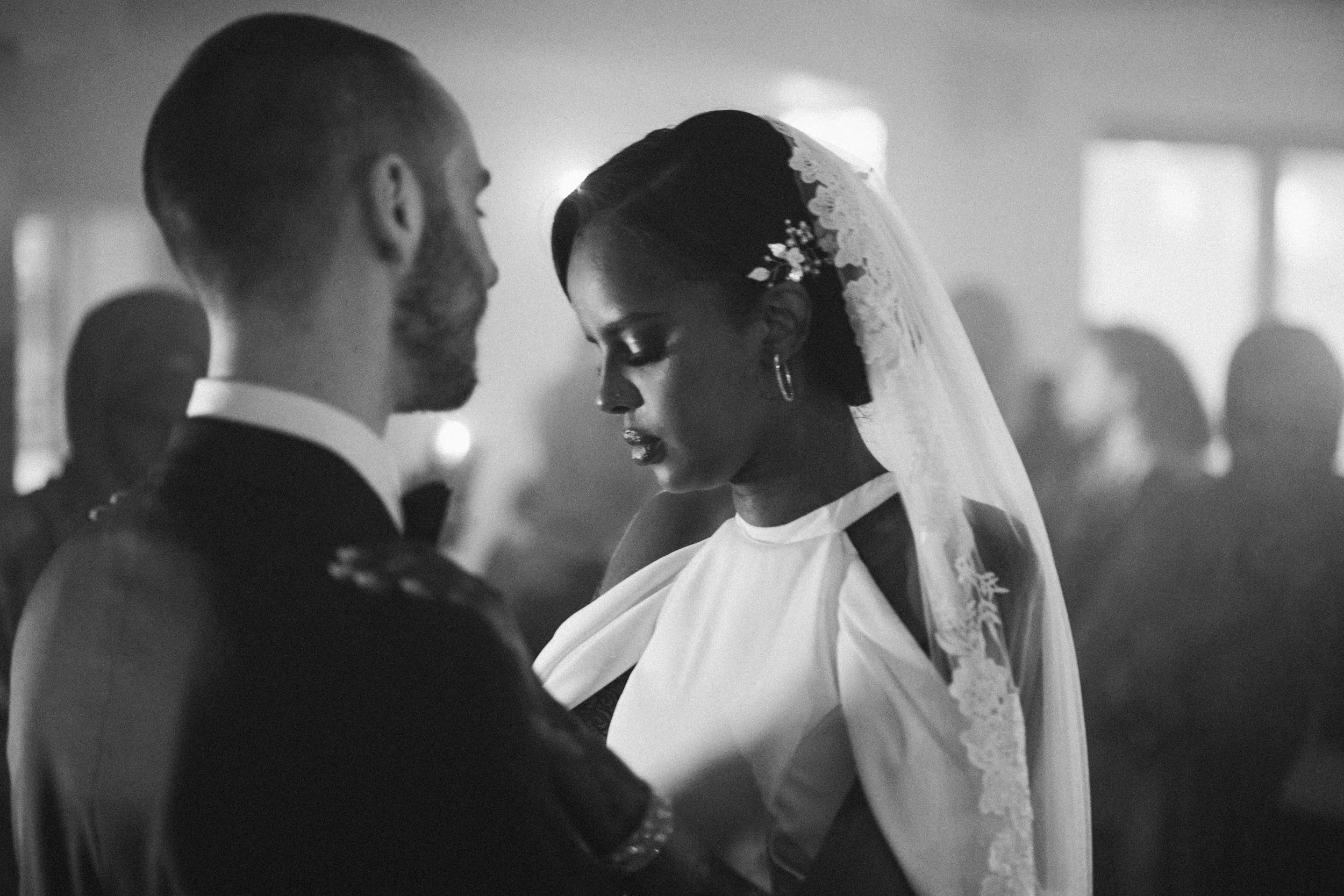 A couple sharing a dance at their wedding reception, with the bride wearing a veil and dress, and the groom in a tuxedo, in a softly lit indoor setting.