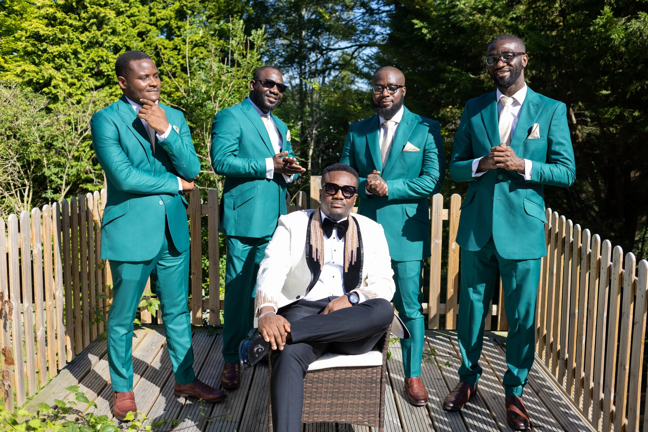 Group of five men dressed in teal suits, one man seated in a white tuxedo with a bow tie, outdoors on a wooden deck with greenery in the background.
