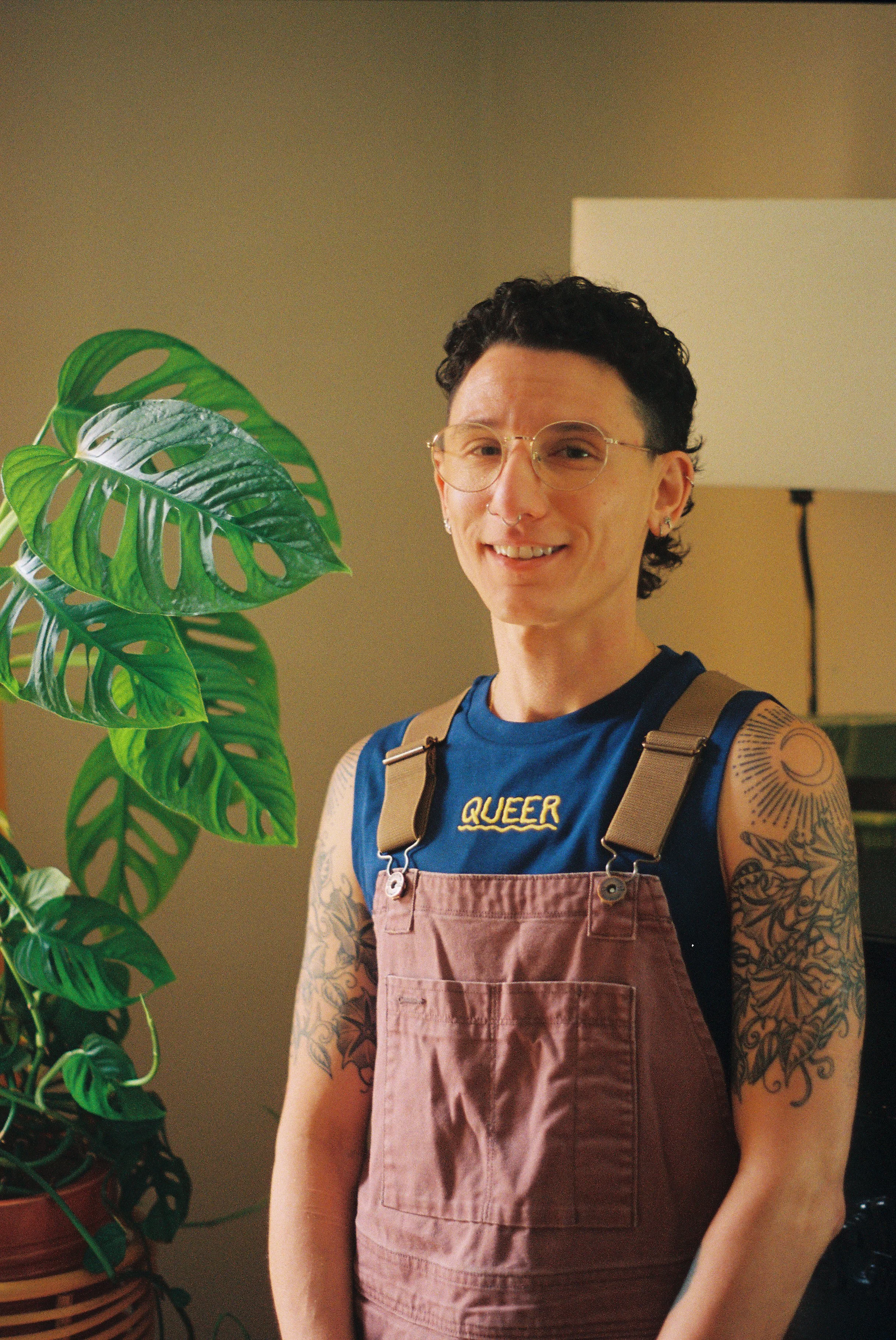 A queer osychologist with short, curly black hair and glasses, wearing a sleeveless shirt with the word 'QUEER' embroidered on the chest and tattoos on their arms, standing indoors next to a large leafy green plant and a lamp in the background.