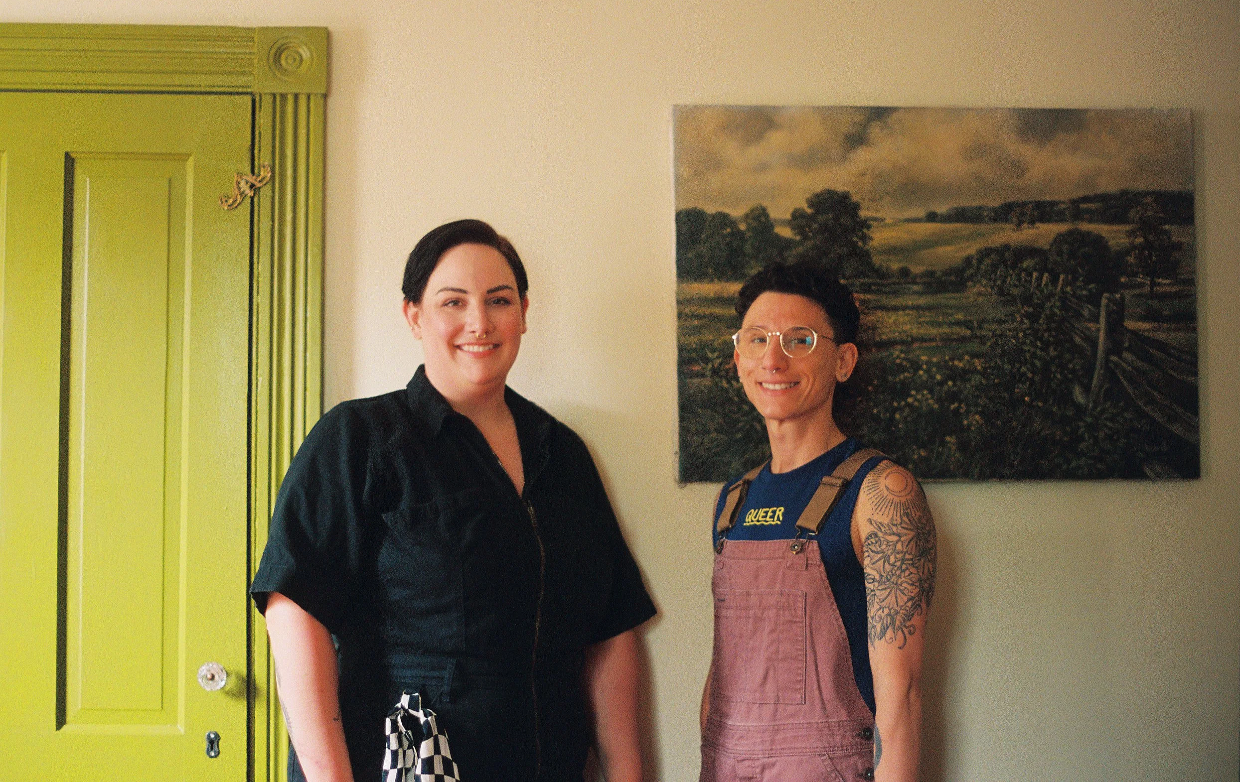 Portrait of two queer psychologists standing indoors, smiling at the camera, with a landscape painting on the wall behind them.