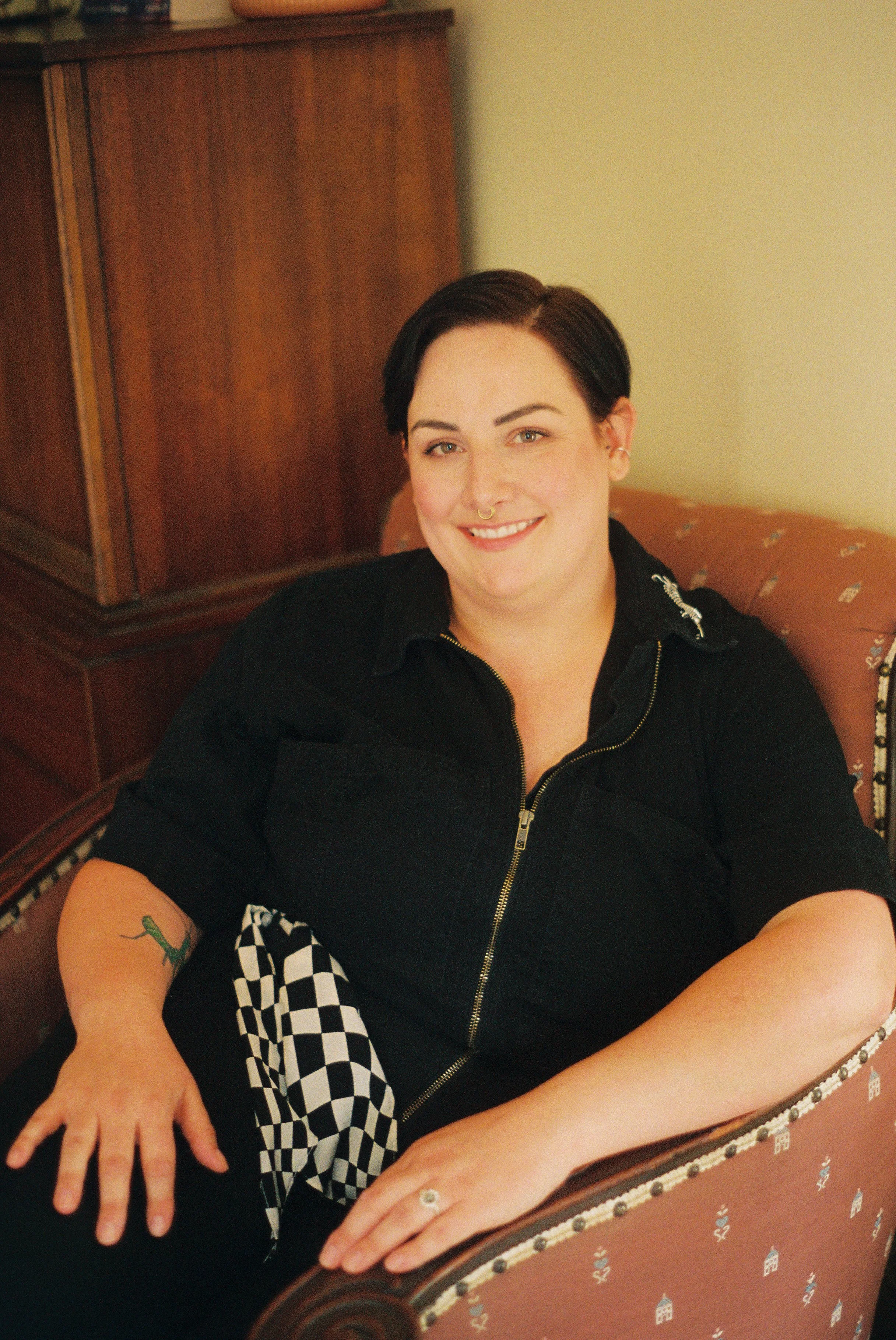 A queer psychologi with short dark hair, wearing a black zip-up jumpsuit, seated on a patterned chair with floral and heart designs, smiling at the camera. They have a septum piercing and a tattoo of an insect on their left arm.