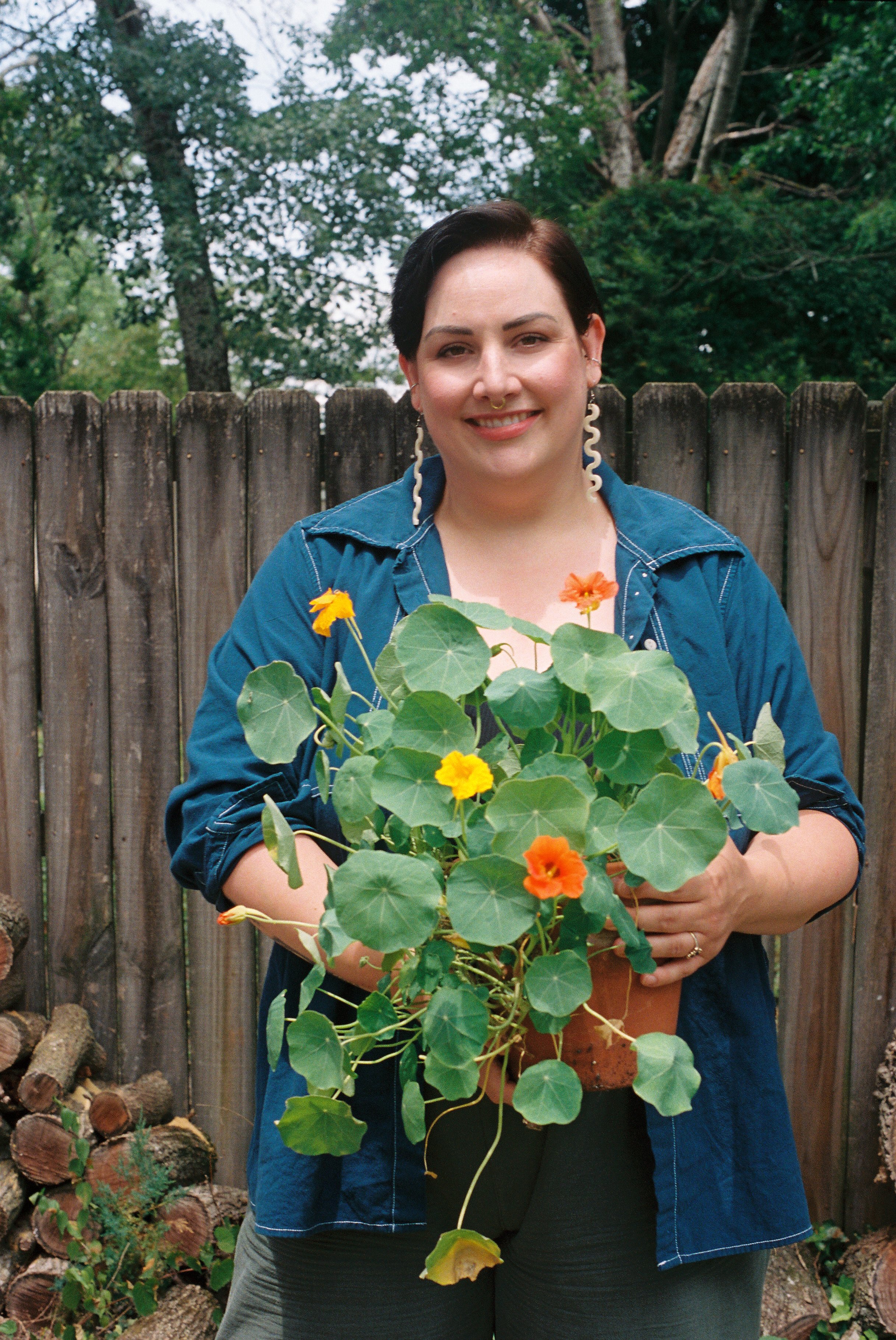A queer psychologist with short black hair, wearing a blue jacket and holding a plant with large green leaves and orange flowers, standing in front of a wooden fence and trees.