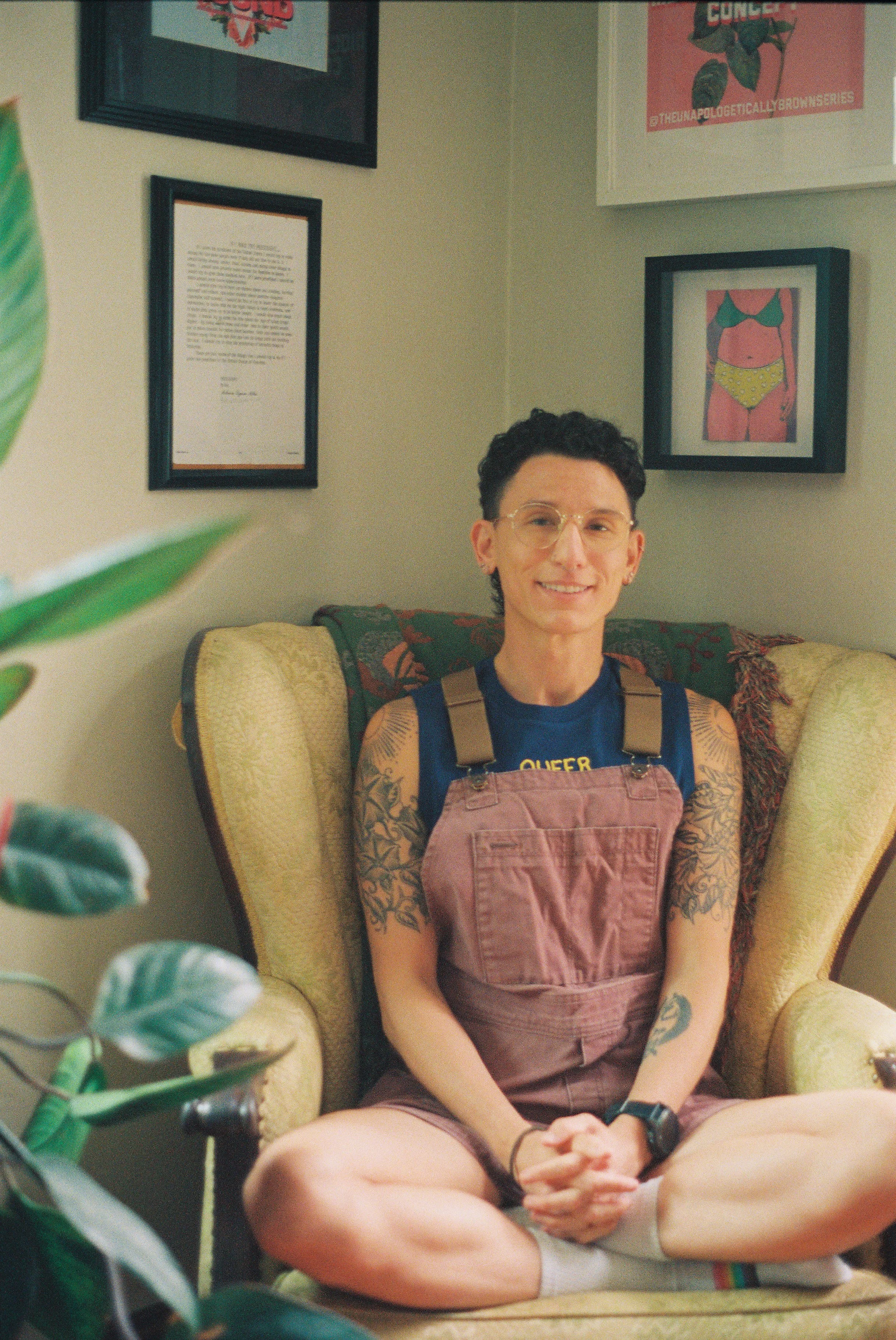 A smiling person with short curly dark hair, glasses, tattoos on arms, wearing a blue T-shirt with yellow text, pink overalls, sitting cross-legged on a yellow armchair with a patterned cushion. The background has framed artwork and posters on beige walls, with some plant leaves visible in the foreground.