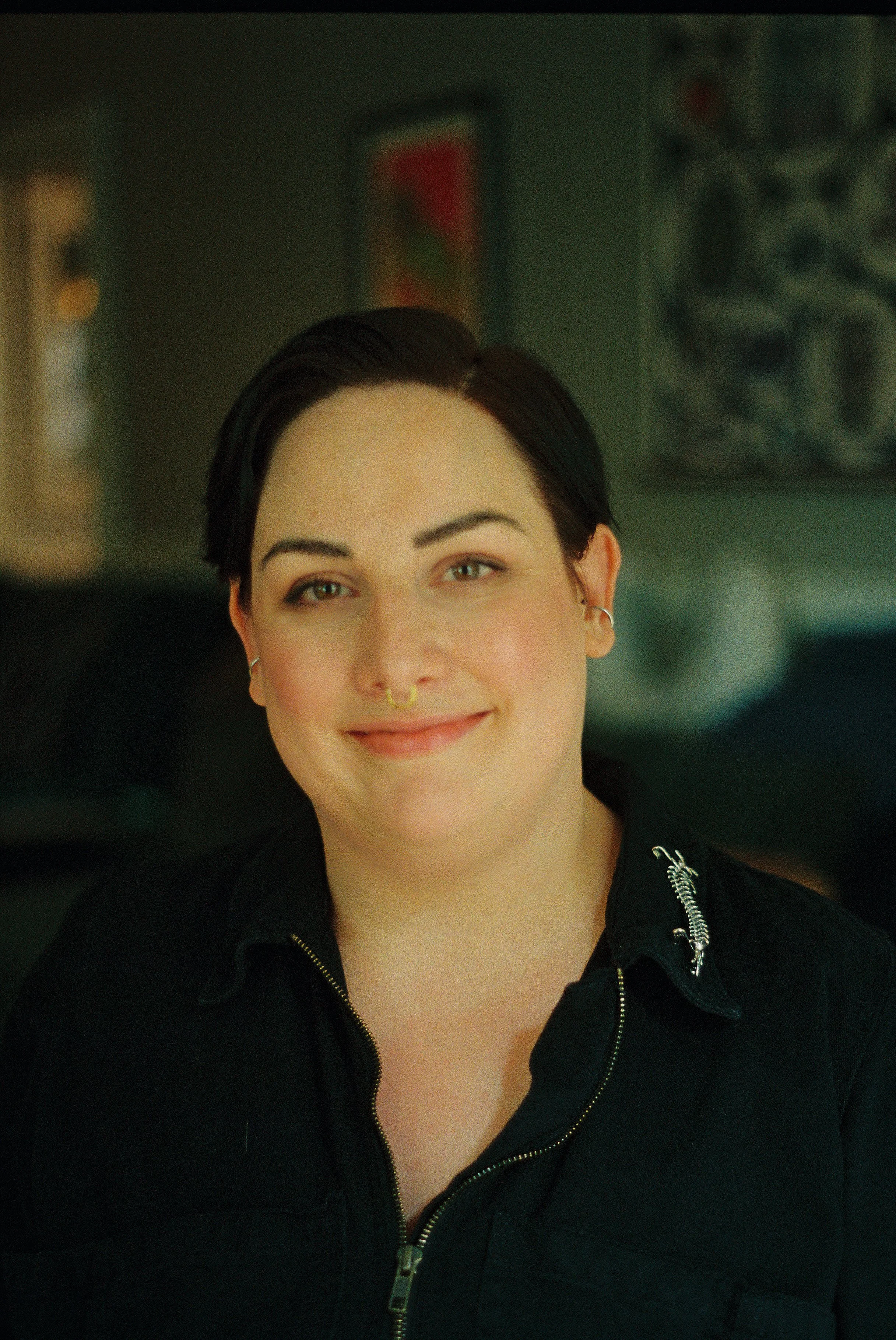 Close-up of a smiling queer psychologist with short dark hair, wearing a septum ring and earrings, dressed in a black jacket with a zipper, in an indoor setting with paintings or posters on the wall in the background.