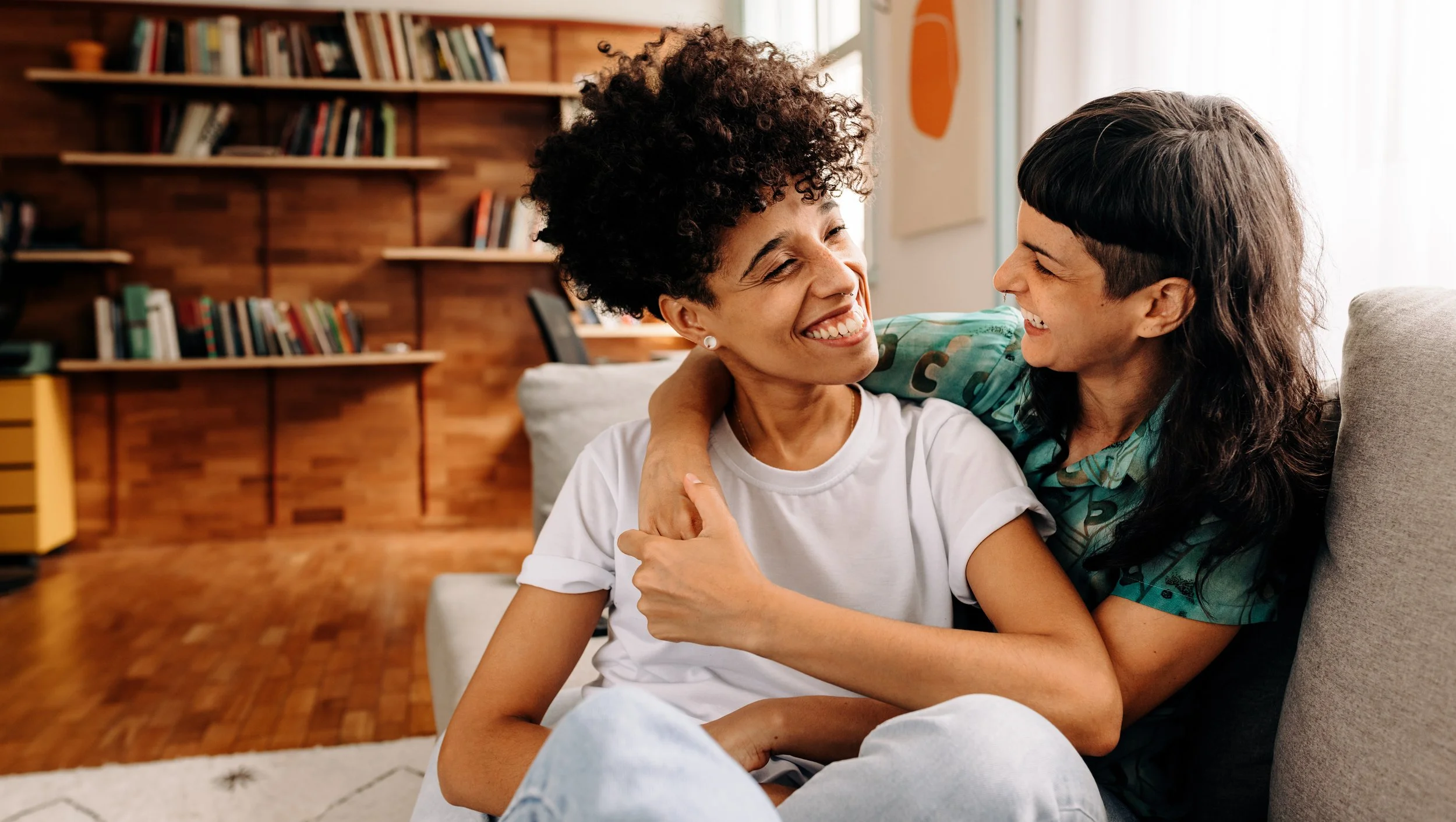 Two queer people, one with curly hair and one with straight black hair, smiling and laughing while sitting on a couch in a cozy living room with a wooden bookshelf in the background.