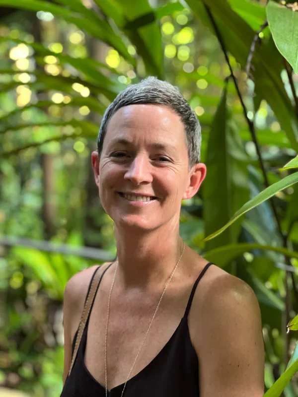 Smiling woman with short gray hair wearing a black tank top and a necklace, standing amidst green tropical plants in a lush outdoor setting.