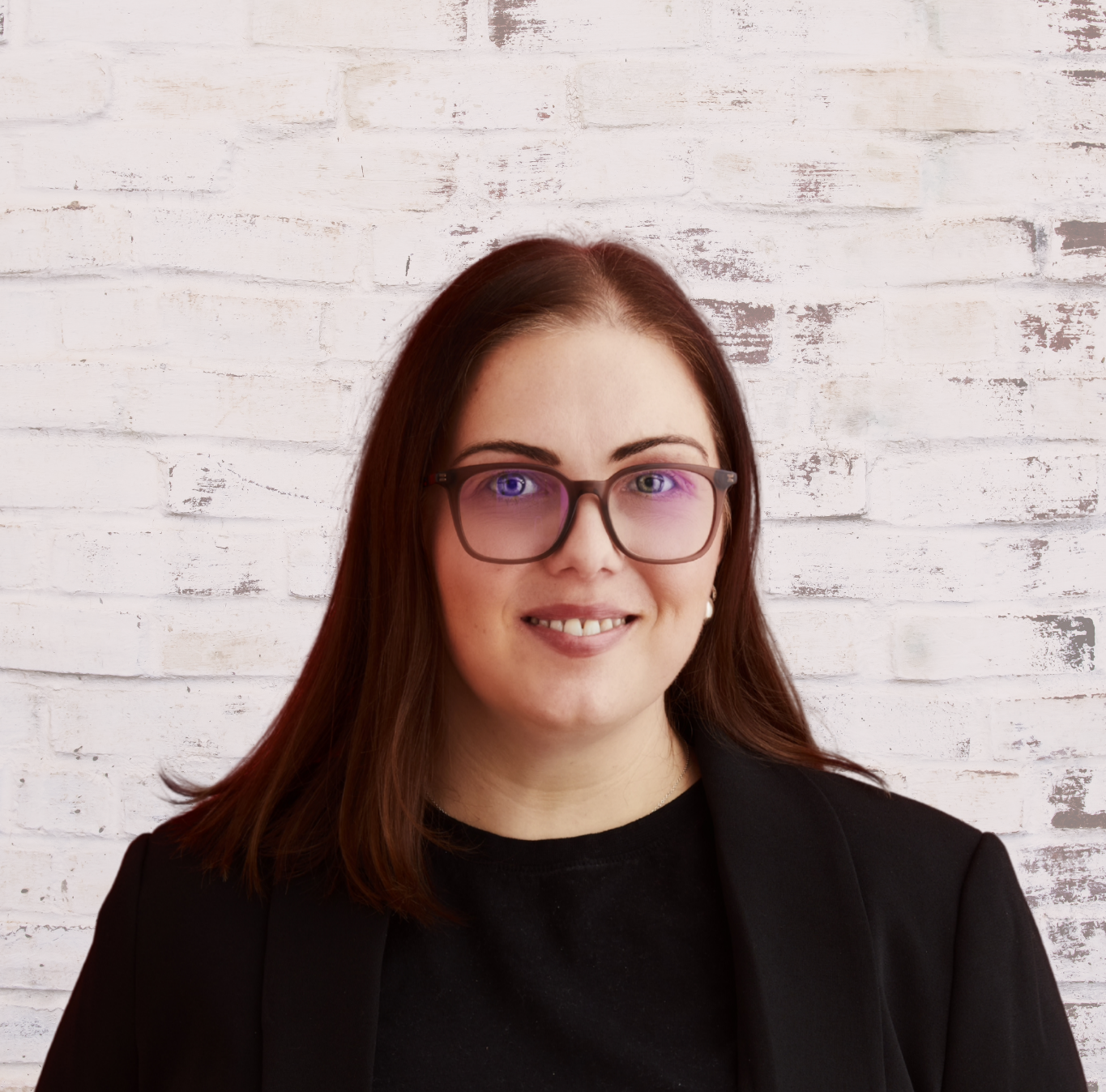 A woman with brown hair, glasses, and a friendly smile, wearing a black blazer, standing in front of a white brick wall.