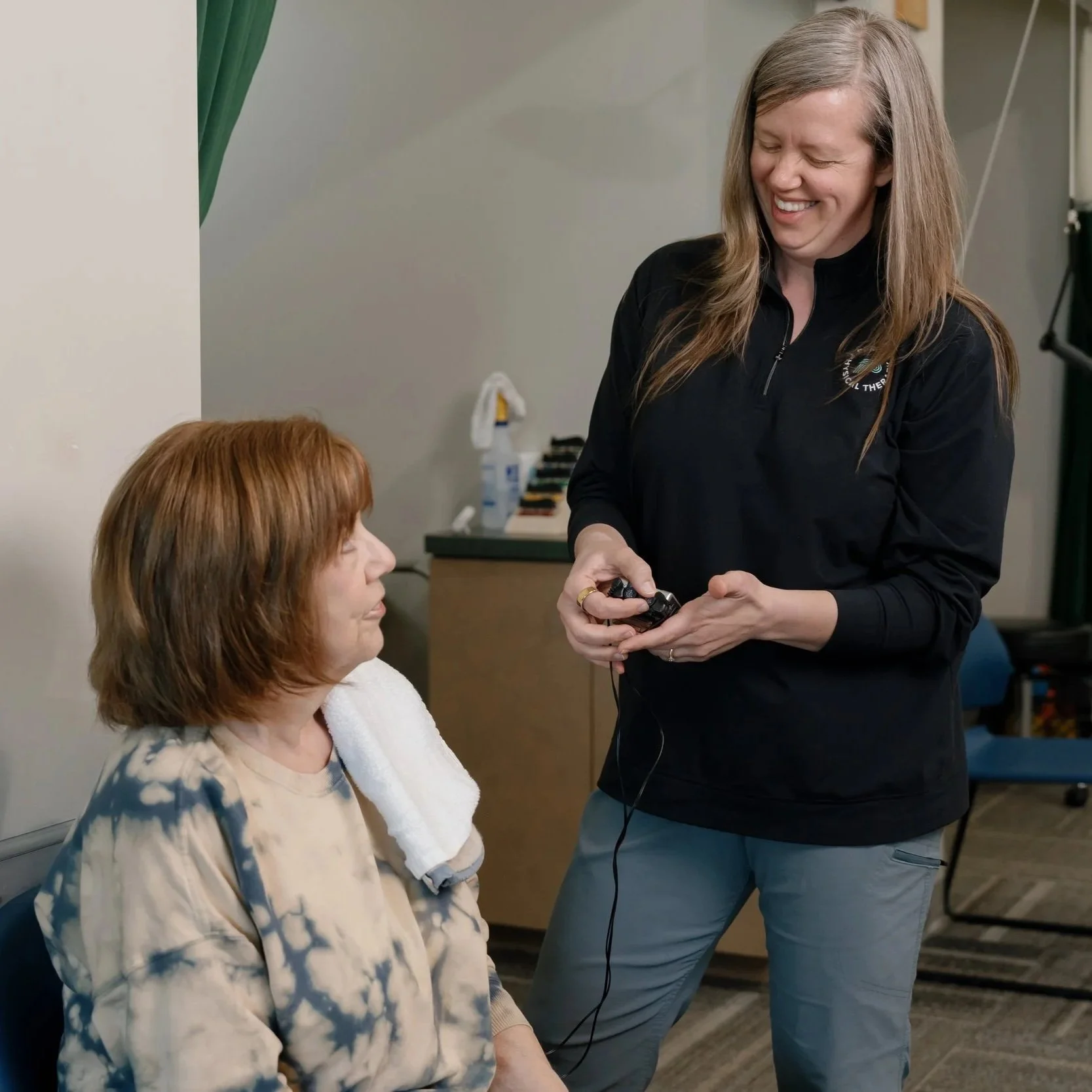 Physical therapist smiling while demonstrating a handheld therapy device to a seated patient in a clinic