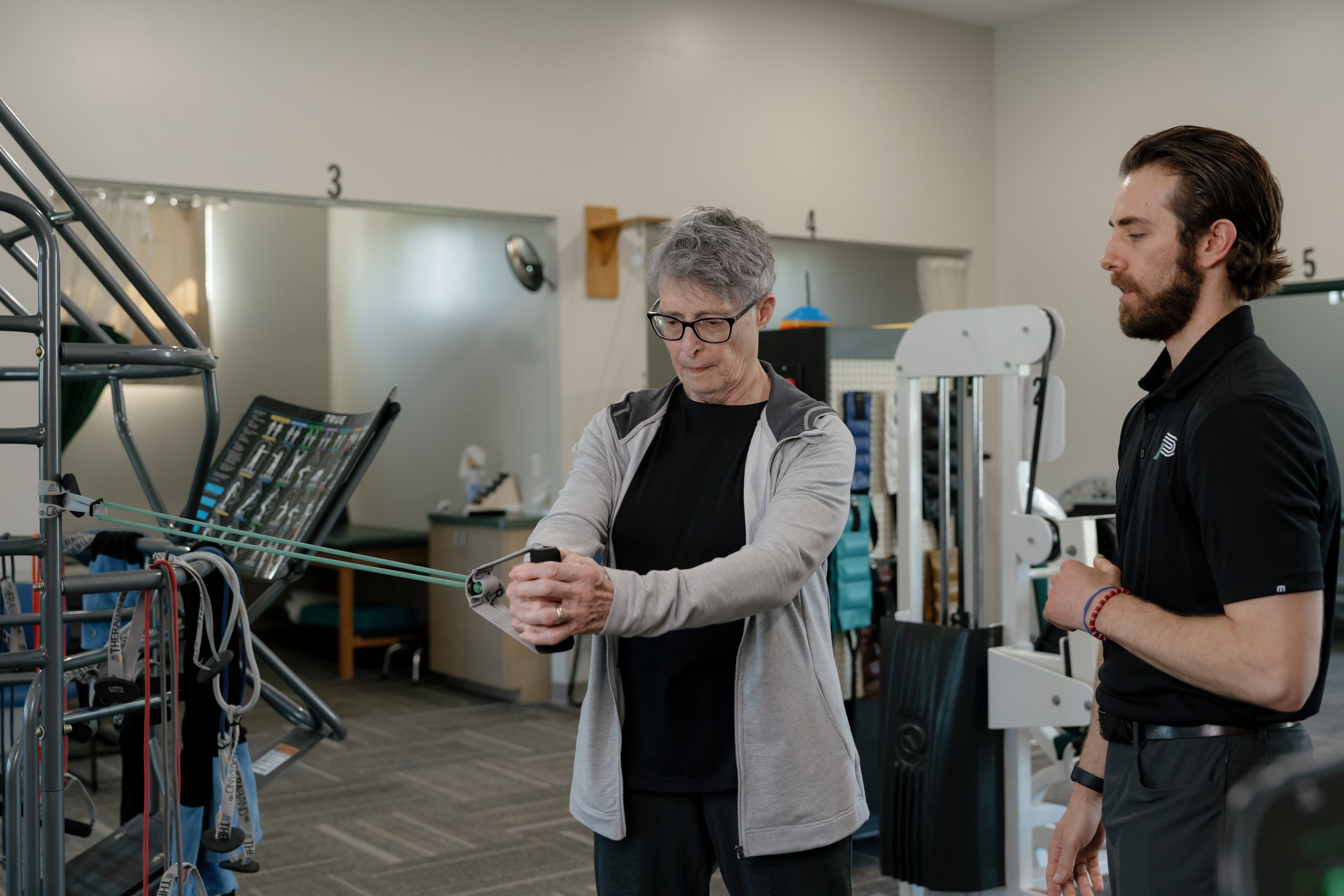Older woman performing resistance-band exercises in a rehabilitation gym while a physical therapist observes.
