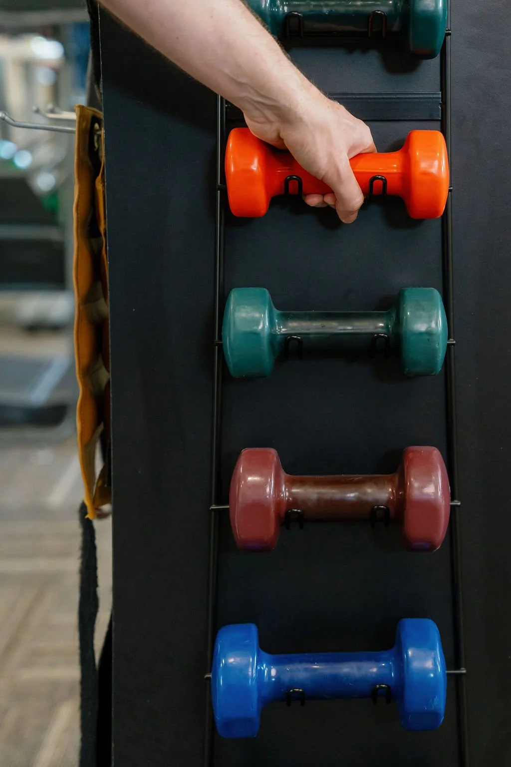Hand removing an orange dumbbell from a vertical rack holding colorful dumbbells in a gym
