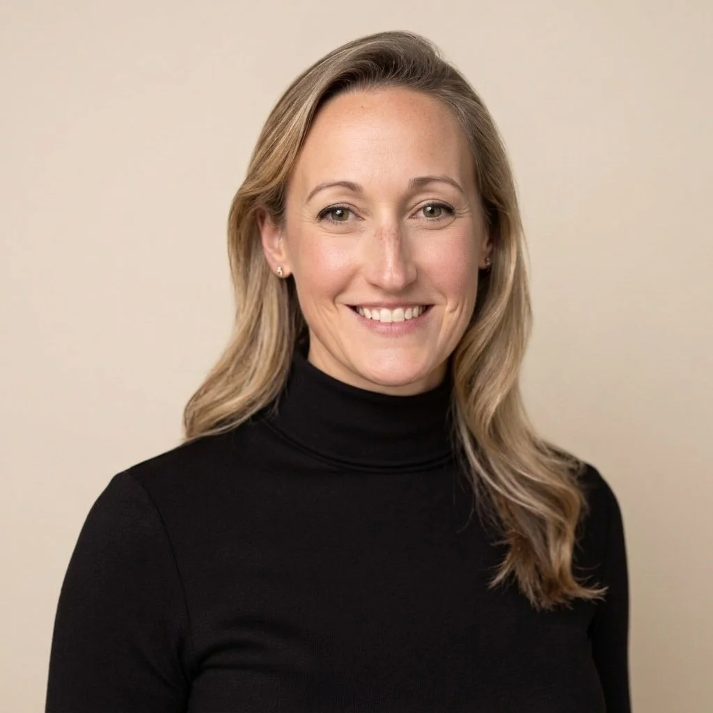 A woman with blonde hair smiling, wearing a black turtleneck, standing against a light neutral background.