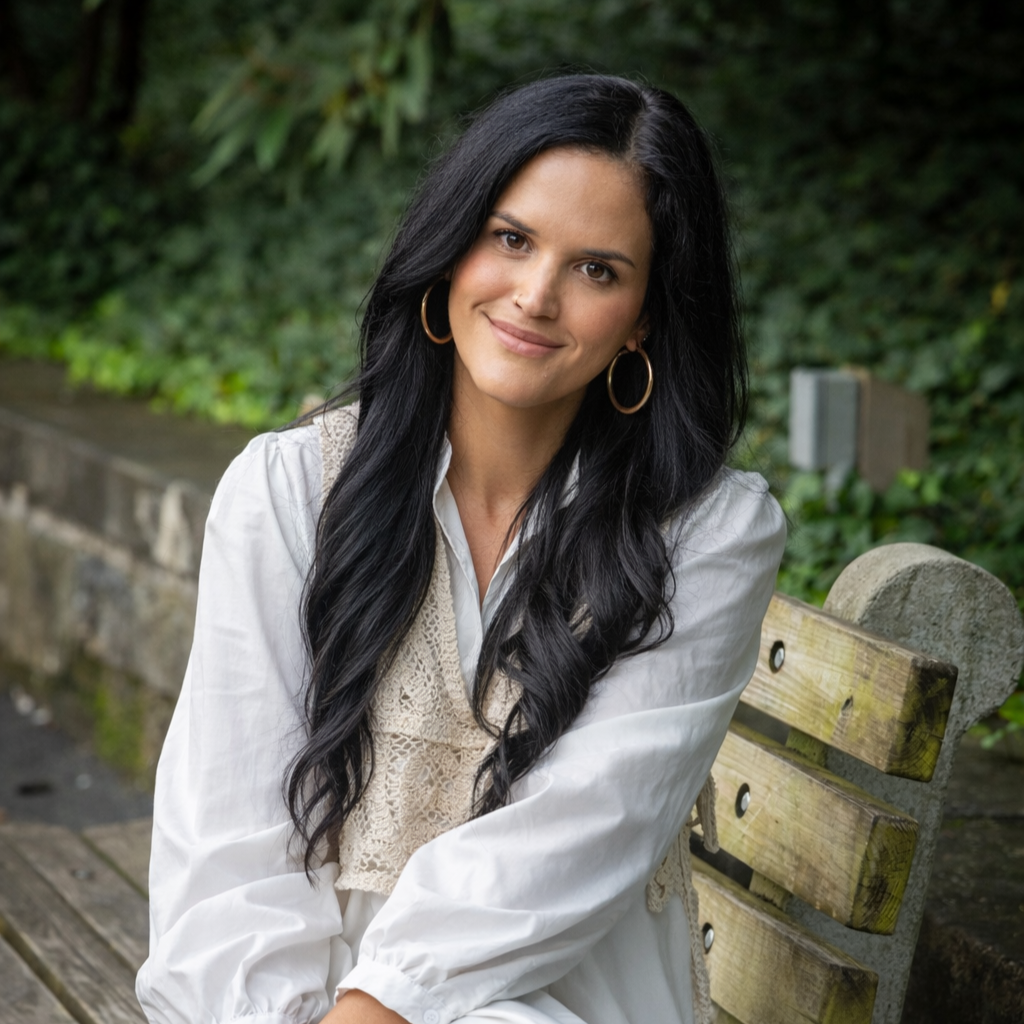 A woman with long black hair, wearing gold hoop earrings and a white blouse with lace details, sitting on a wooden bench outdoors surrounded by greenery.