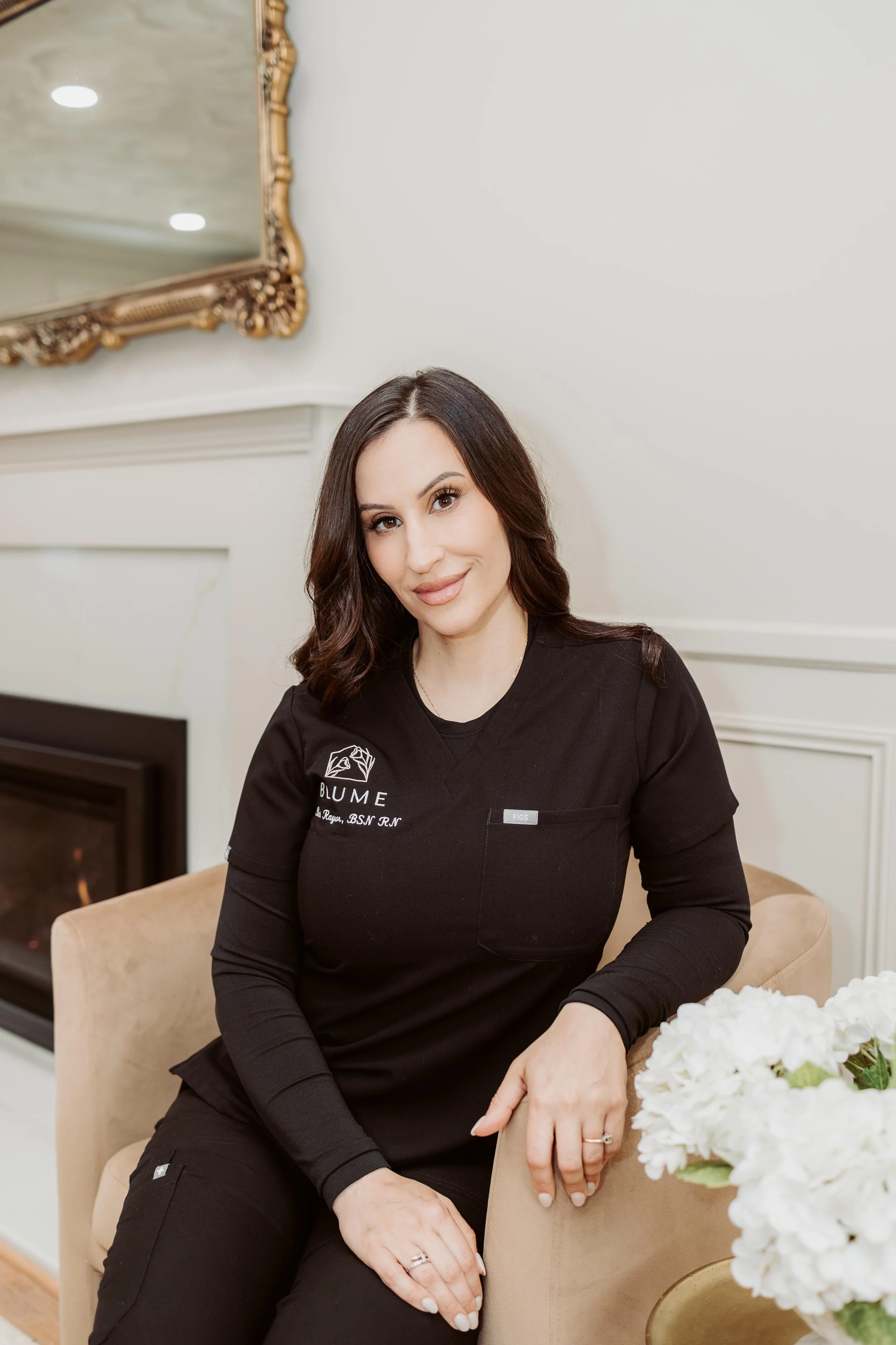 A woman with dark hair wearing a black long-sleeve top, sitting on a beige chair in a room with cream walls and a large ornate mirror reflected in the background.
