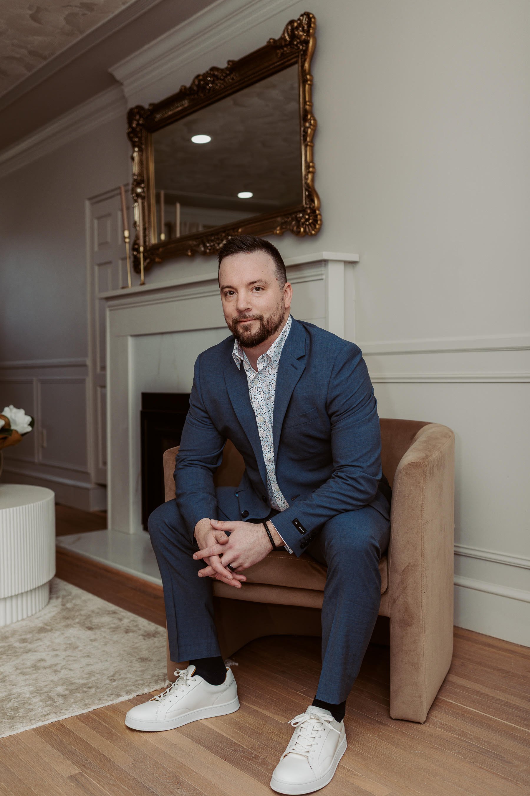 A man in a blue suit and white sneakers sitting on a beige chair in a living room with a fireplace and ornate mirror.