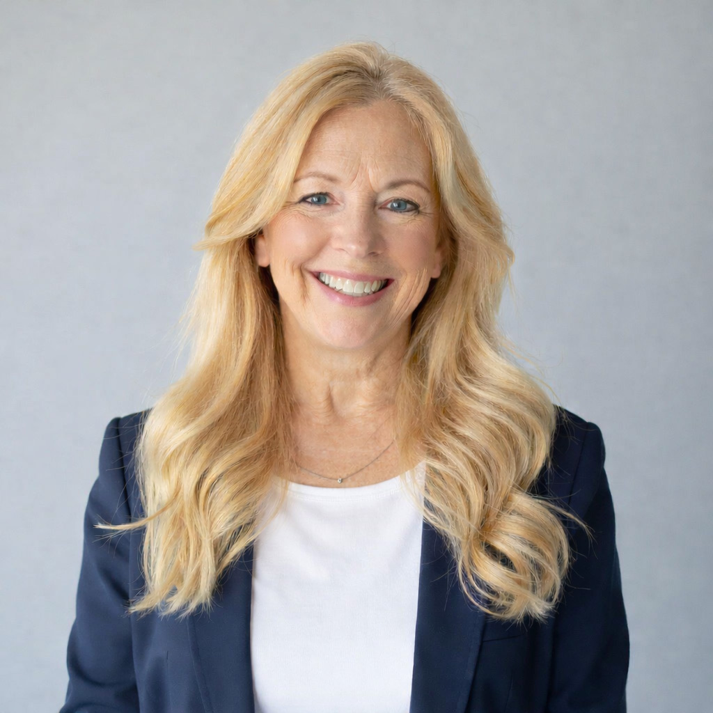 A smiling woman with long blonde hair wearing a navy blazer and a white shirt, standing against a light grey background.