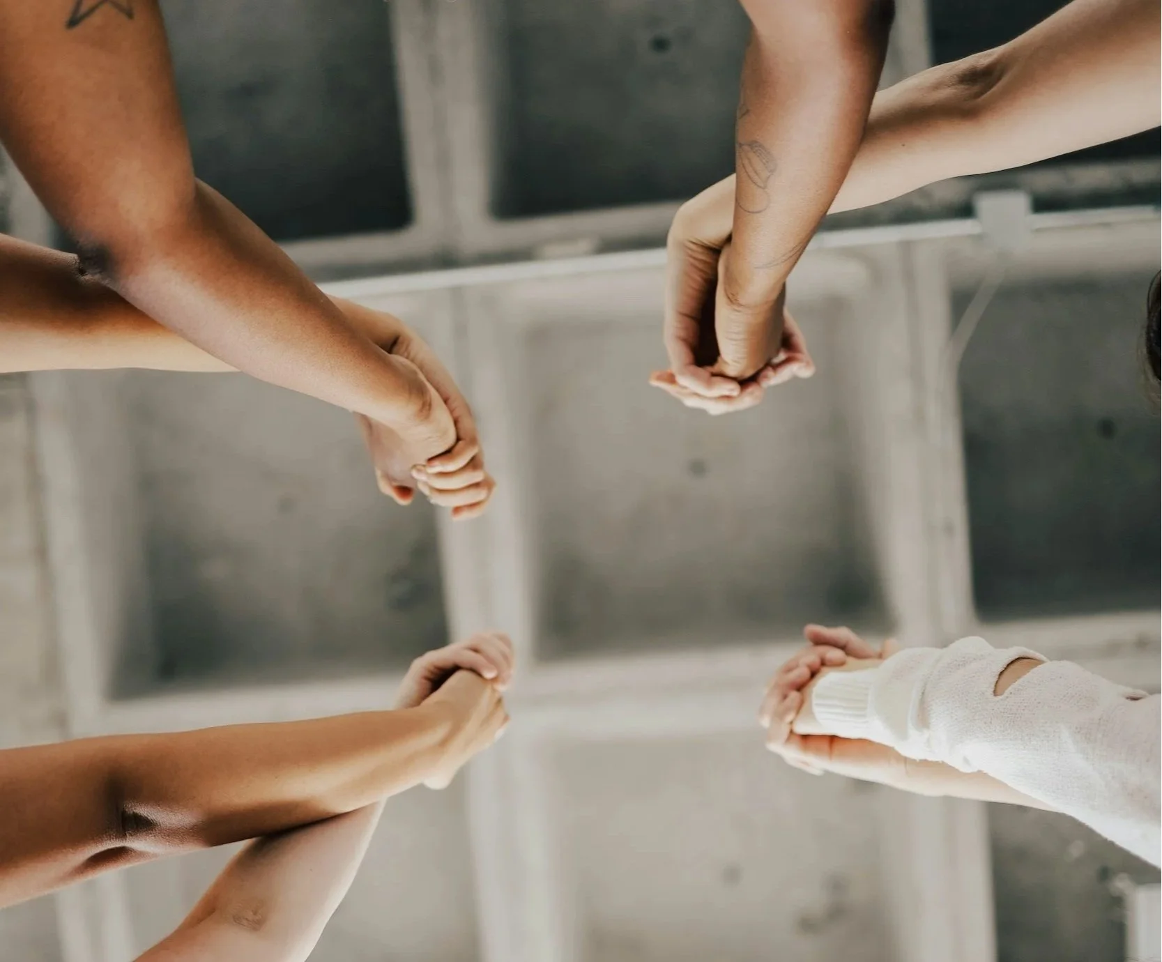 Multiple people's hands holding each other's fingers in a circle, shot from above with a stainless steel sink in the background.