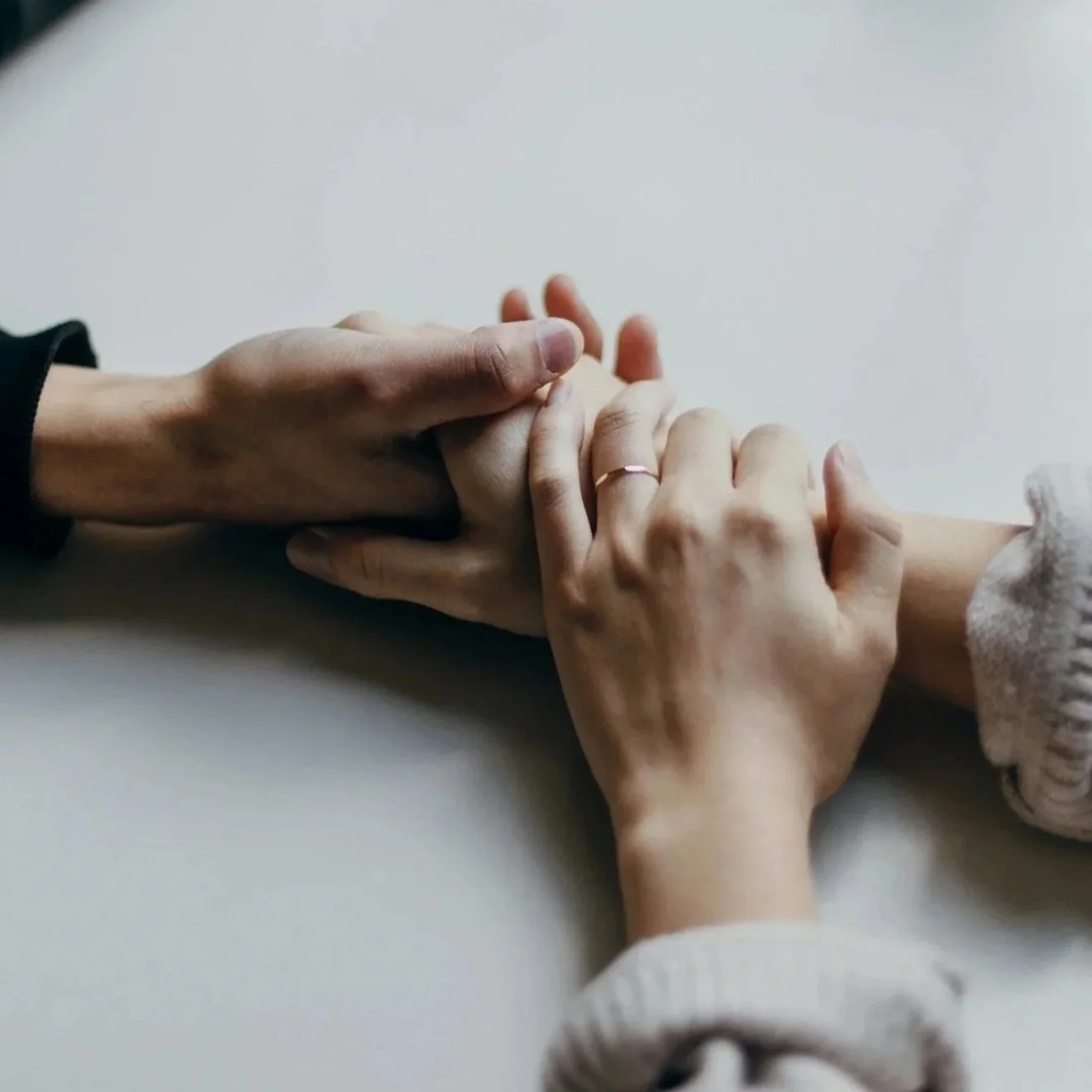 Two hands gently holding each other's fingers on a plain white surface, one hand wearing a ring and a long-sleeve shirt.