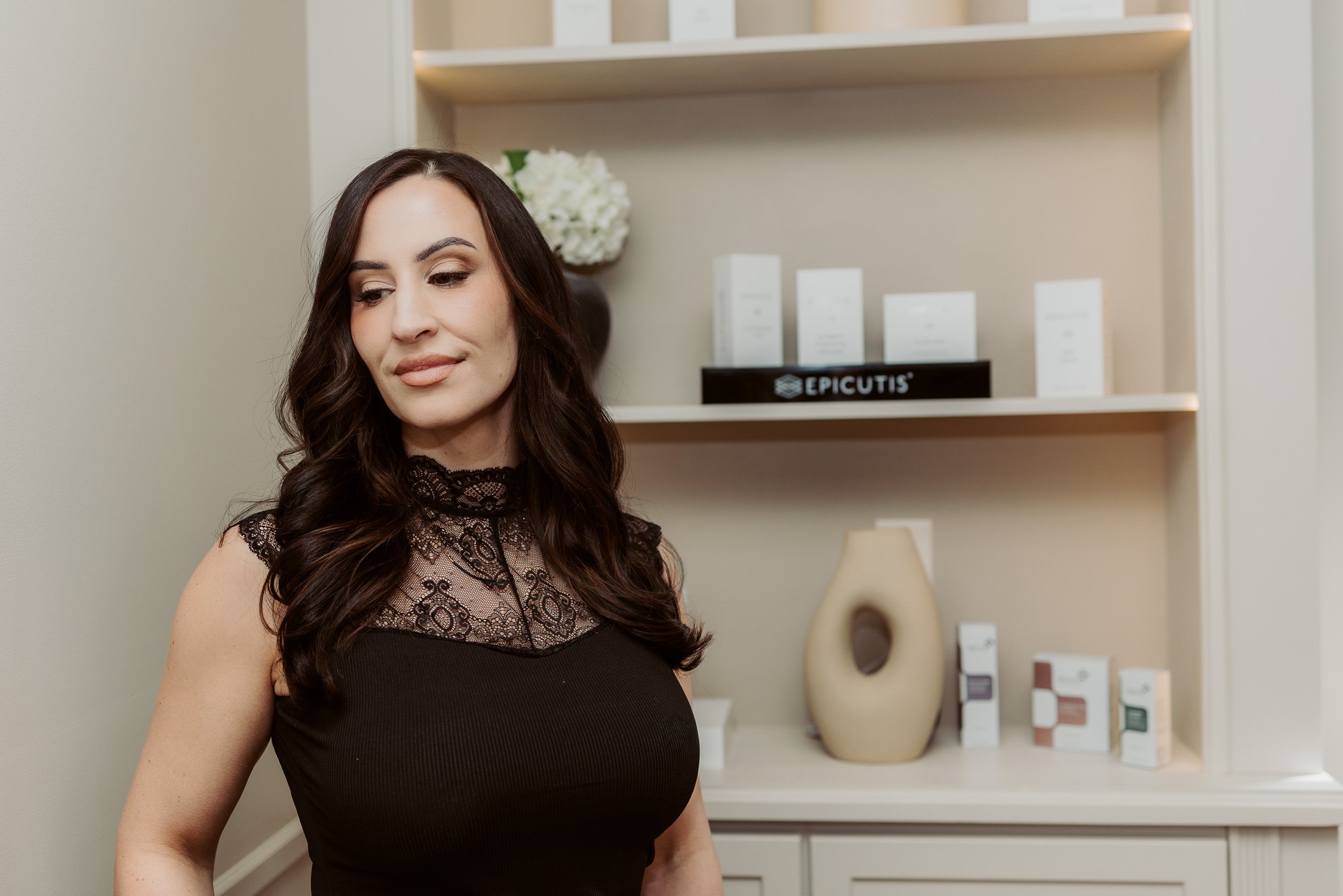 A woman with long dark hair and in a black lace top stands indoors near a shelving unit with white boxes and decorative items.