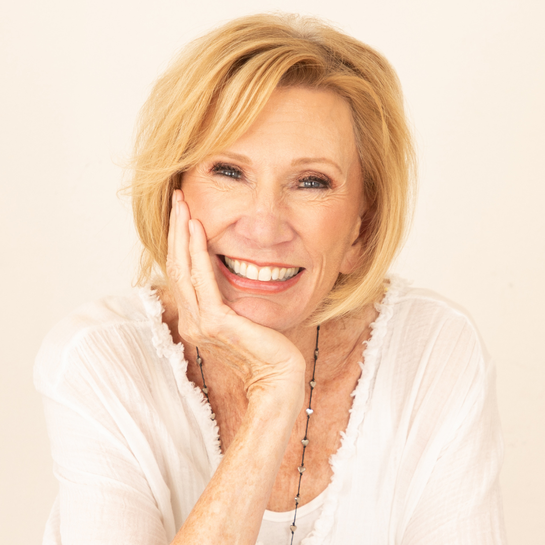 A smiling middle-aged woman with blonde hair, resting her face on her hand, wearing a white top and a necklace.