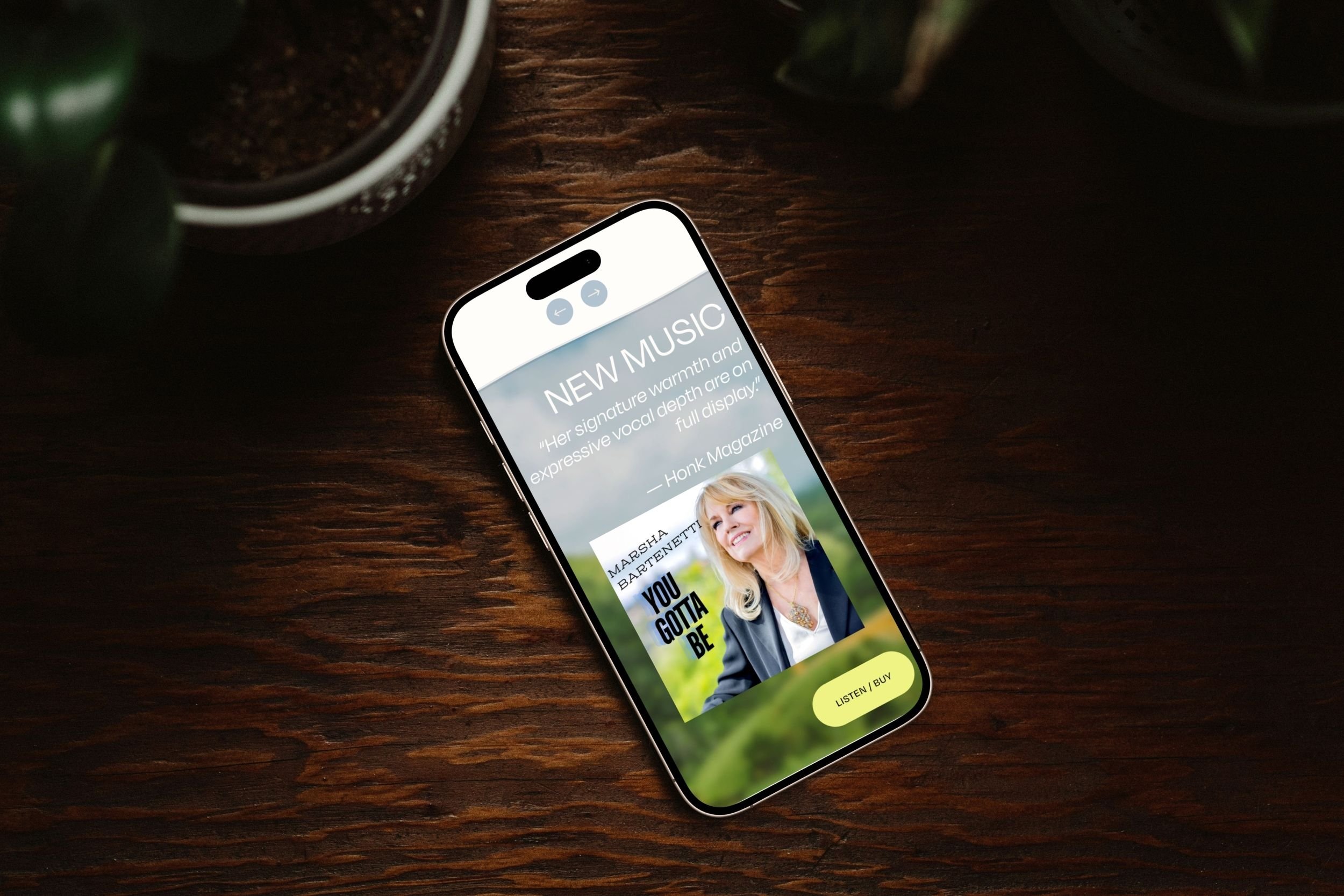A smartphone on a wooden table displaying a magazine cover featuring Marsha Bartnetti with text "You Gotta Be" and buttons for listening or buying. Part of a potted plant is visible in the background.
