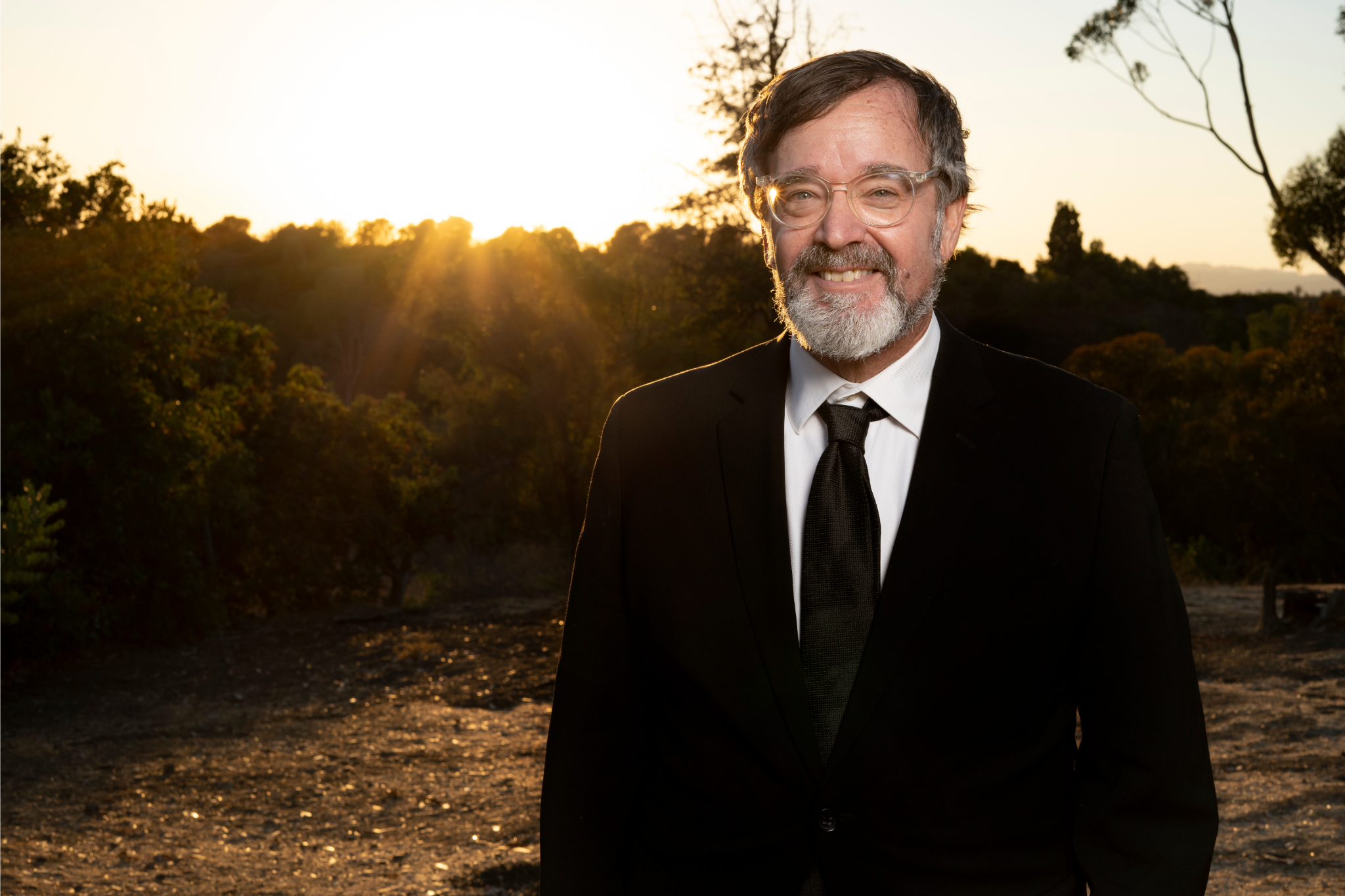 A smiling man with glasses, gray beard, wearing a suit and tie, standing outdoors during sunset with trees and a clear sky in the background.
