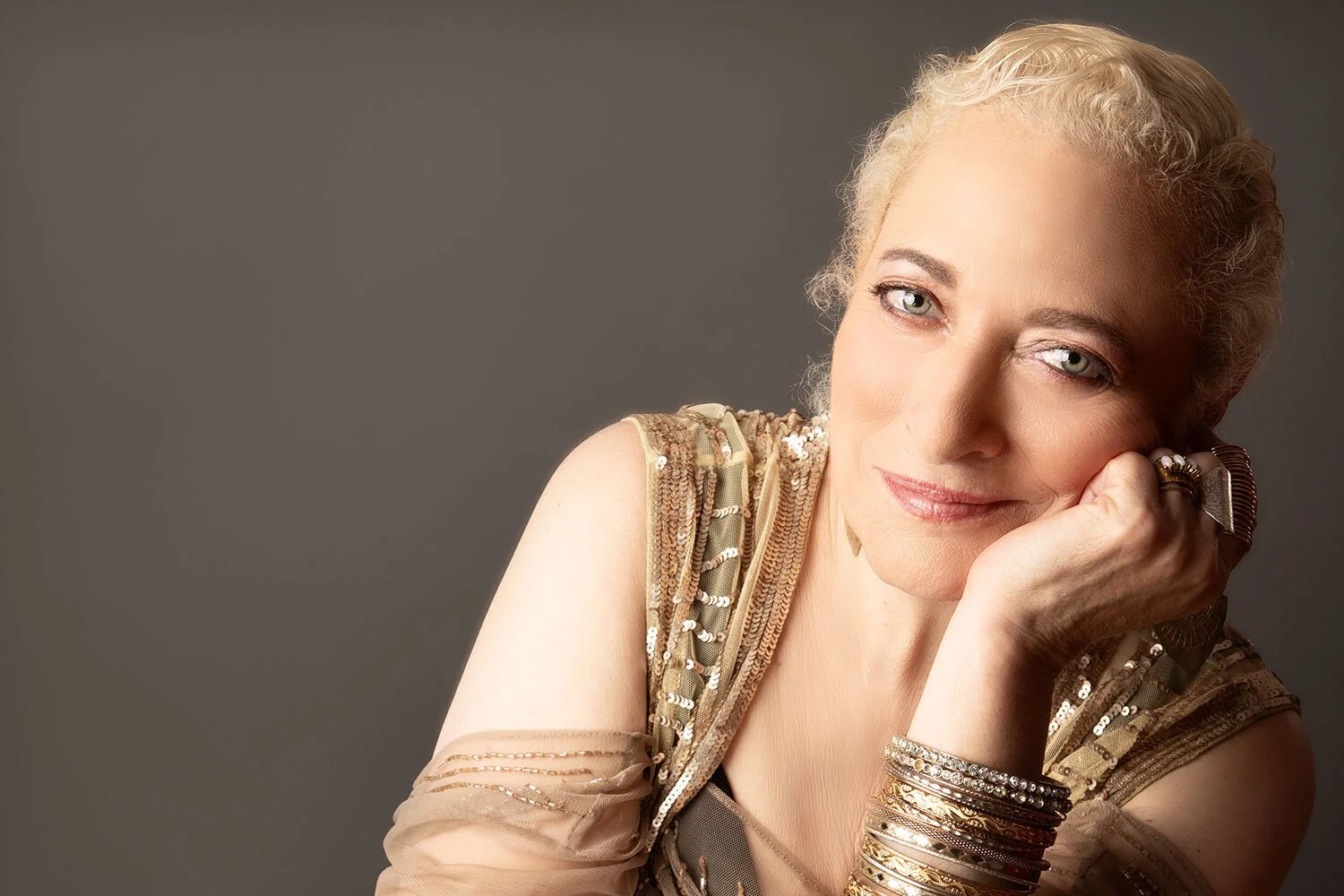 A middle-aged woman with blonde hair styled in soft waves, wearing a gold-sequined sleeveless top, resting her chin on her hand, adorned with multiple gold bangles and rings, smiling softly against a dark gray background.