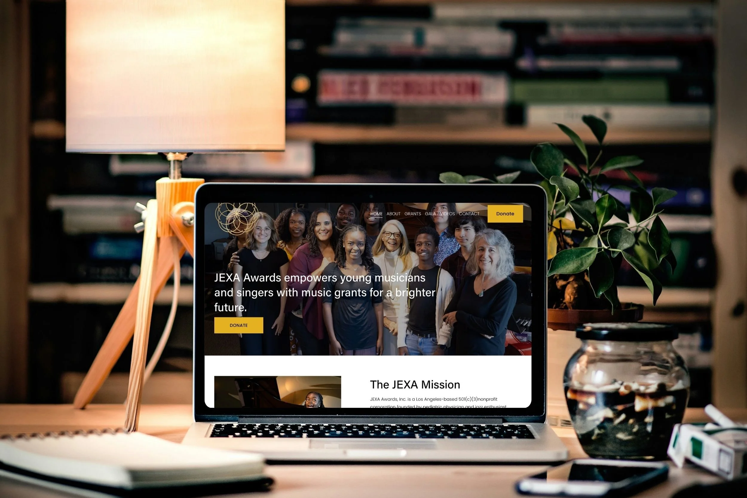 A laptop on a desk displaying a website for JEXA Awards with a photo of a diverse group of young people and text about empowering young musicians with music grants, with a yellow "Donate" button.
